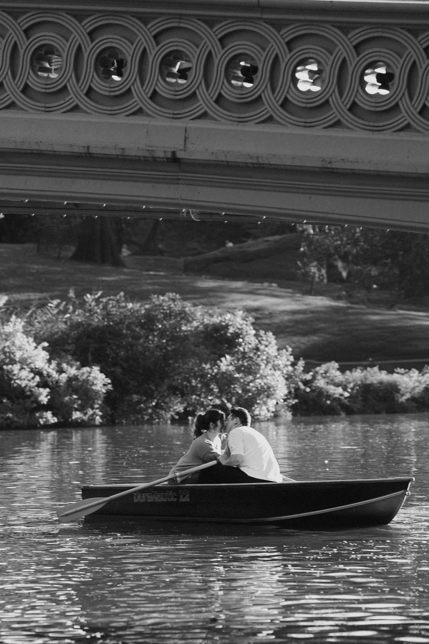 A black and white image of a rowboat on a lake with ornate bridge railing visible above.