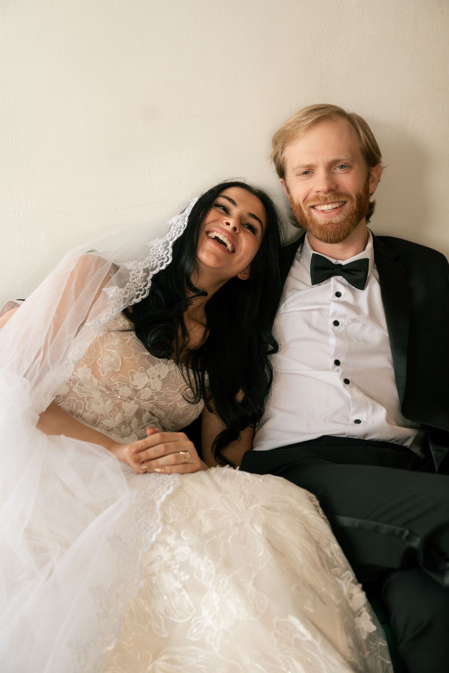 A joyful wedding couple laughs together while sitting against a neutral wall, the bride wearing a lace veil and gown.