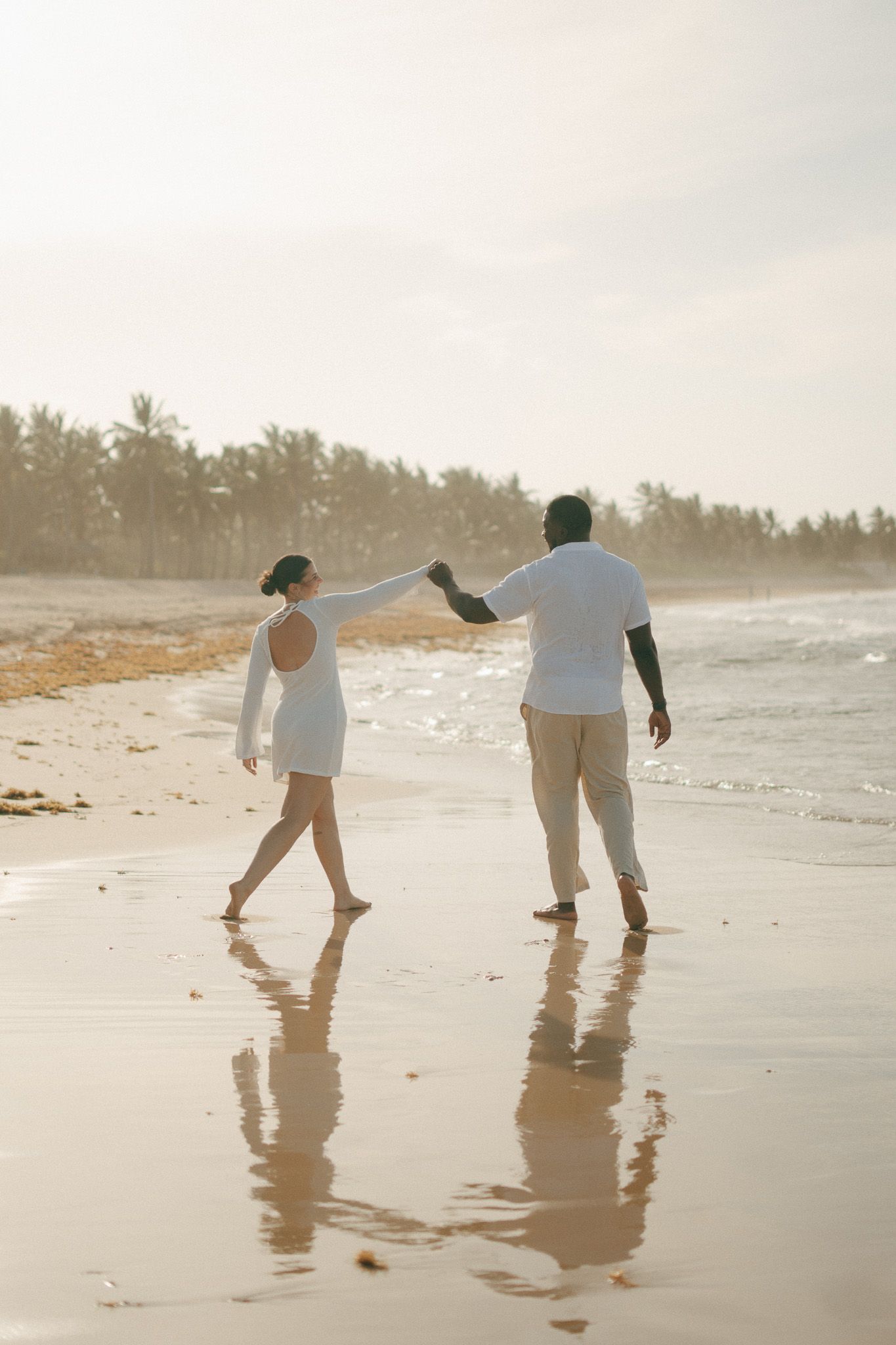 Couple in white outfits holding hands and walking barefoot on a sunny tropical beach with palm trees.