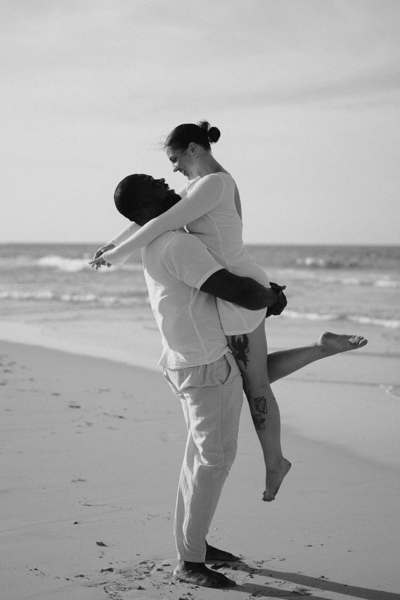 Black and white photo of a man lifting a woman on a beach, both wearing white, sharing a romantic moment by the ocean.
