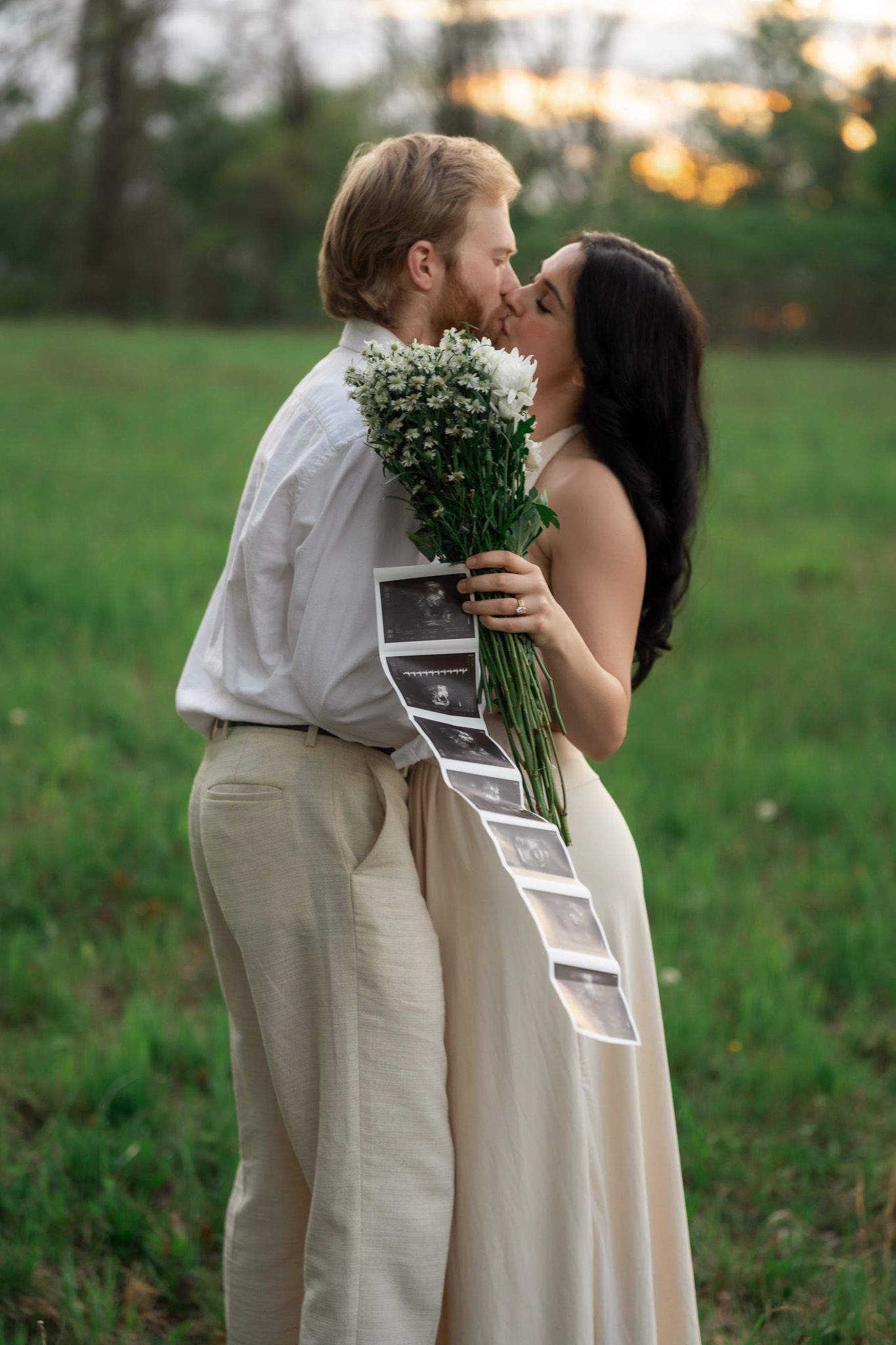 Couple kissing in a green field at sunset, holding white flowers and ultrasound photos, announcing pregnancy.