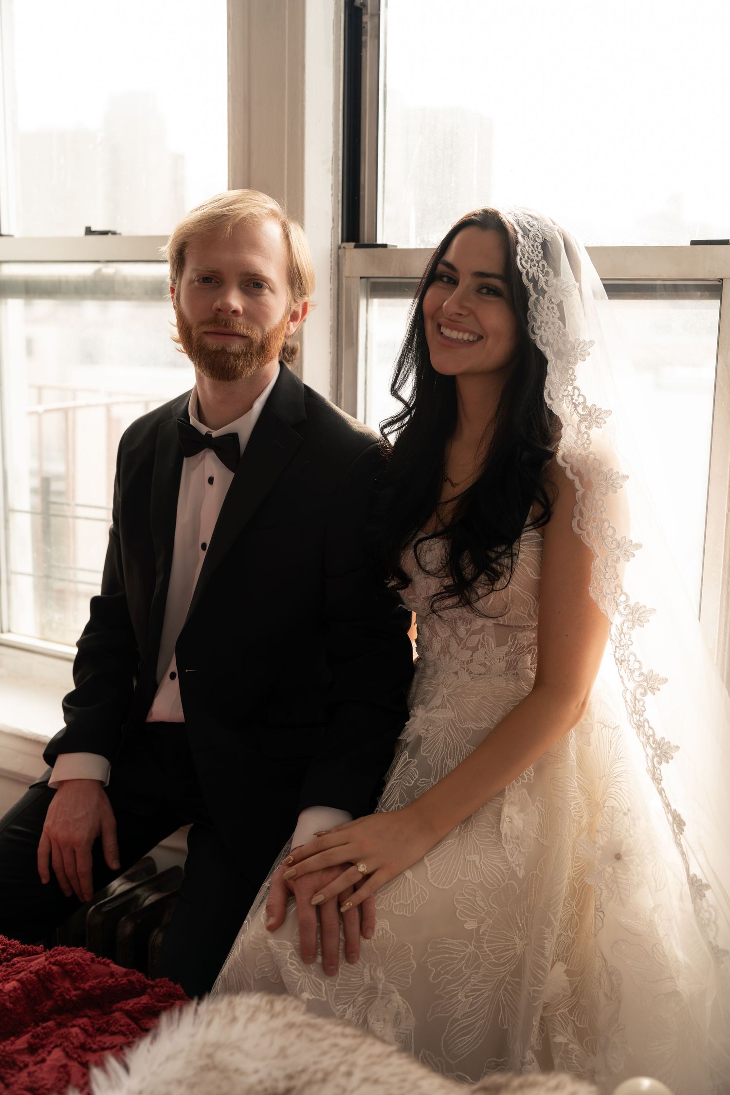 A couple poses together by a window on their wedding day, with the bride wearing a white lace gown.