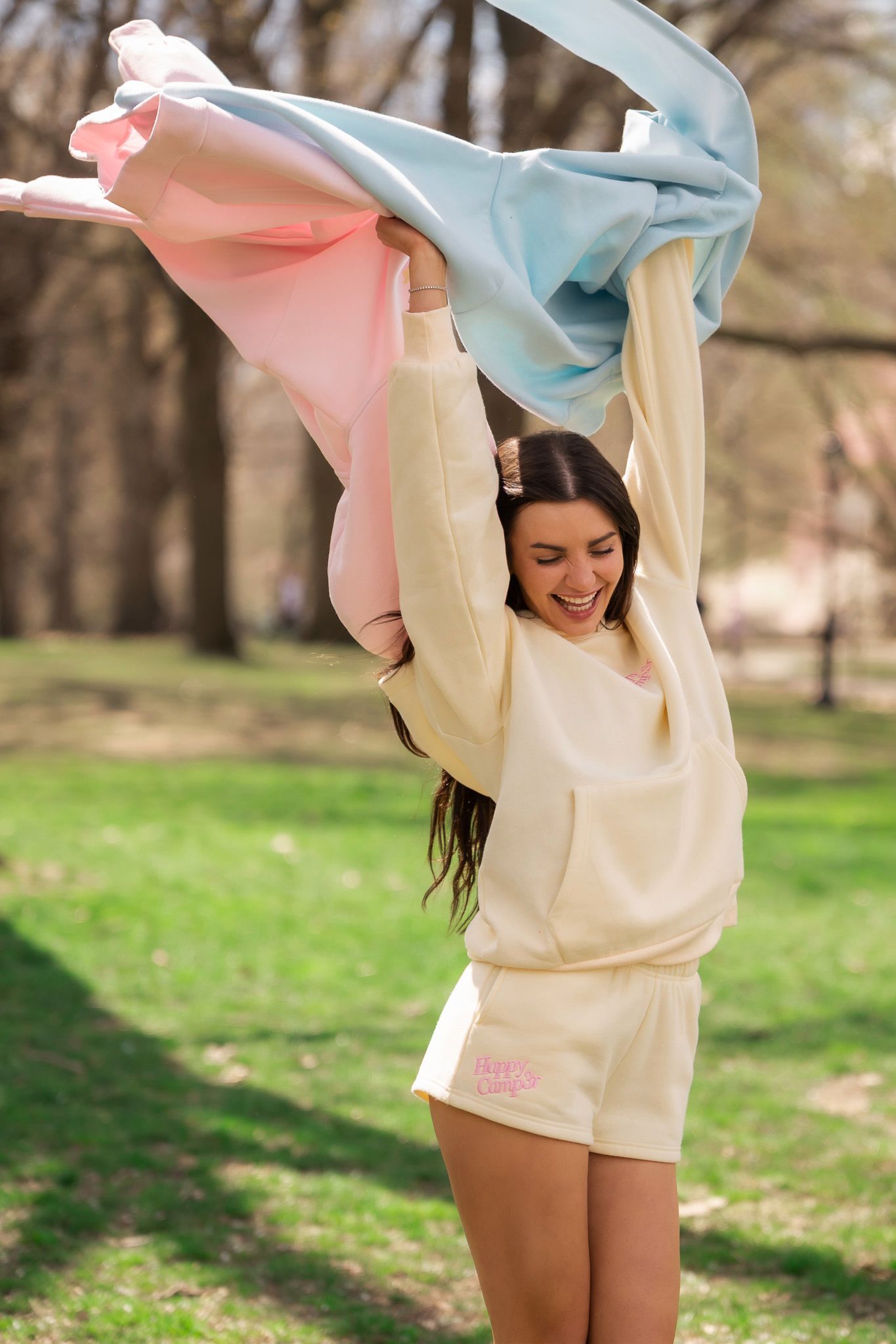 Young woman in cream sweater and shorts joyfully holds colorful pink and blue fabric overhead in a sunny park.