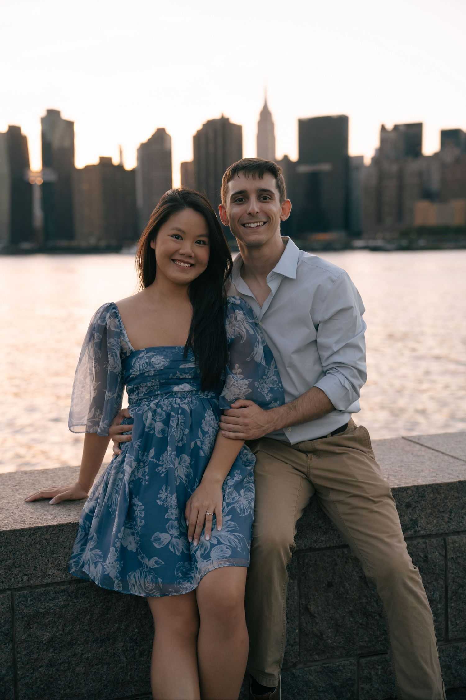A couple sits together on a waterfront wall with the New York City skyline silhouetted in the background at sunset.