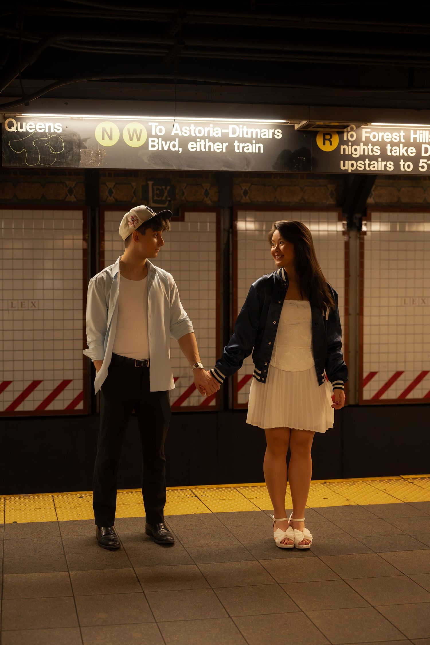Couple holding hands on NYC subway platform near N/W train signs at Queens station.