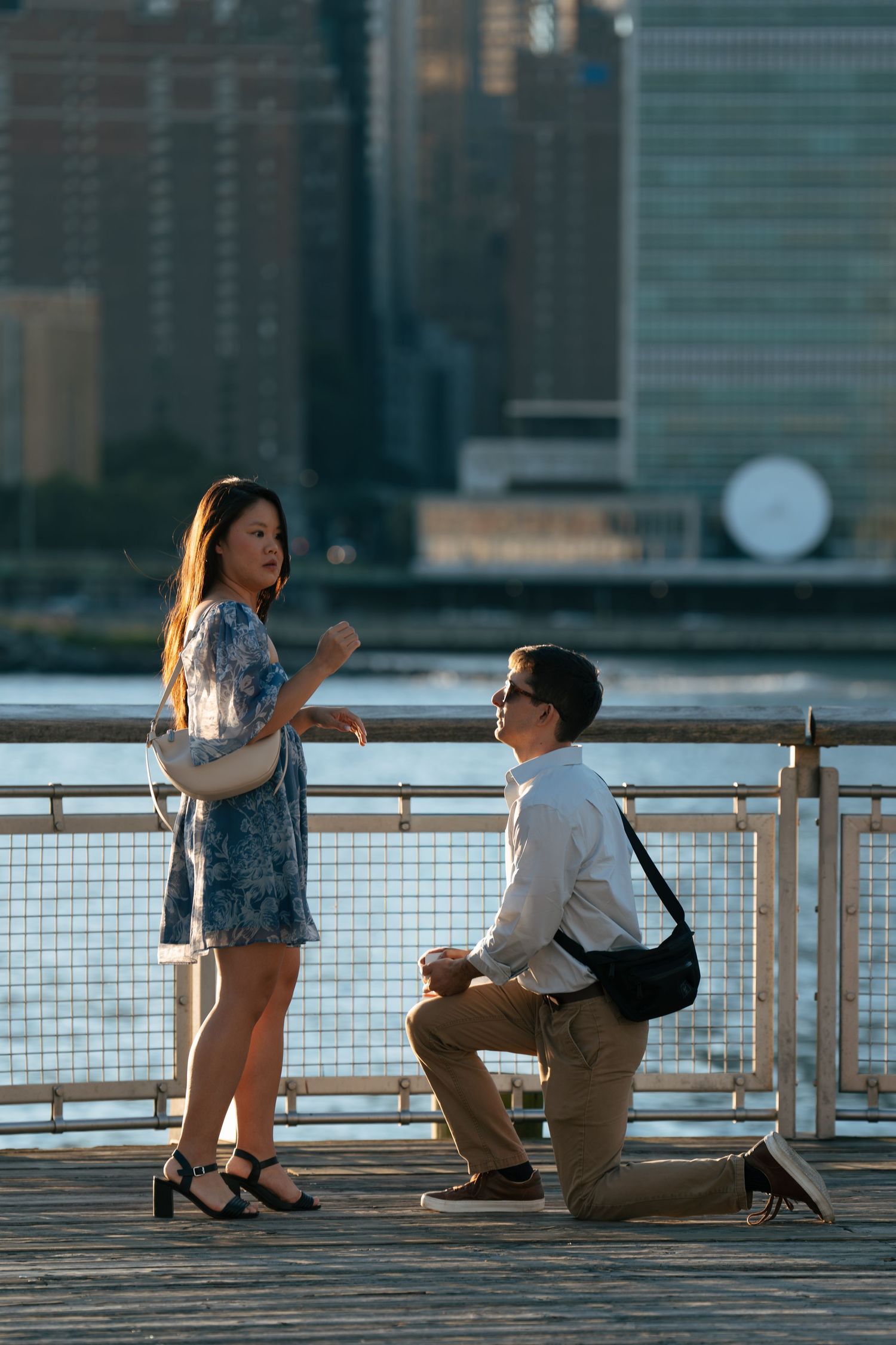 A couple shares a romantic moment during a sunset proposal on a waterfront pier with the city skyline in the background.