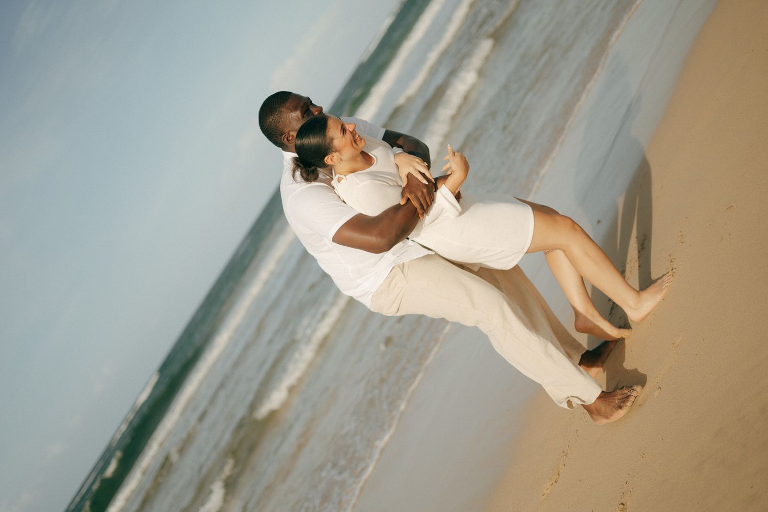 Romantic couple embracing on a sandy beach, dressed in white, with ocean waves in the background.