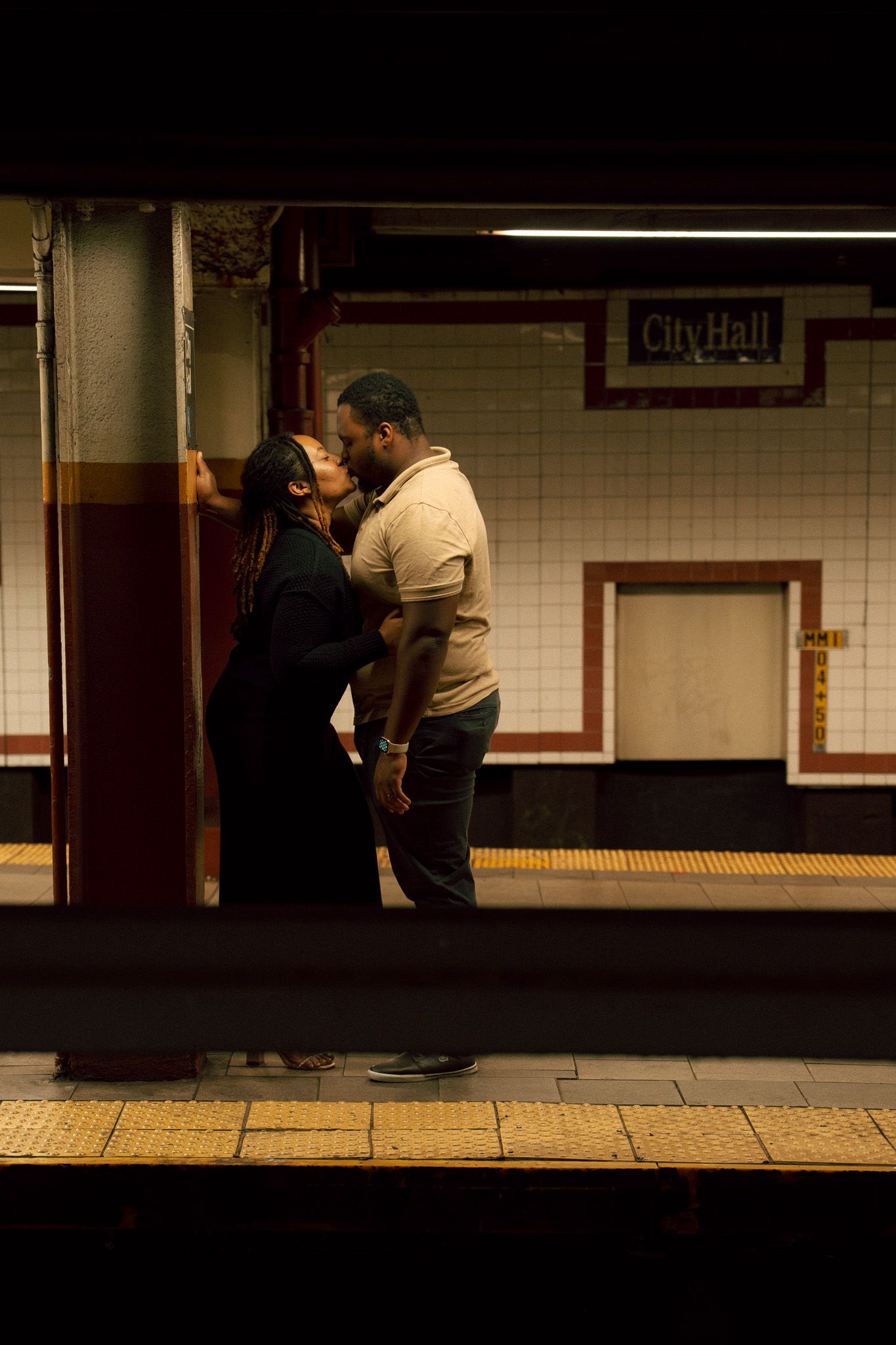 Silhouetted figures stand on a dimly lit subway platform with white tiled walls and yellow safety strip.