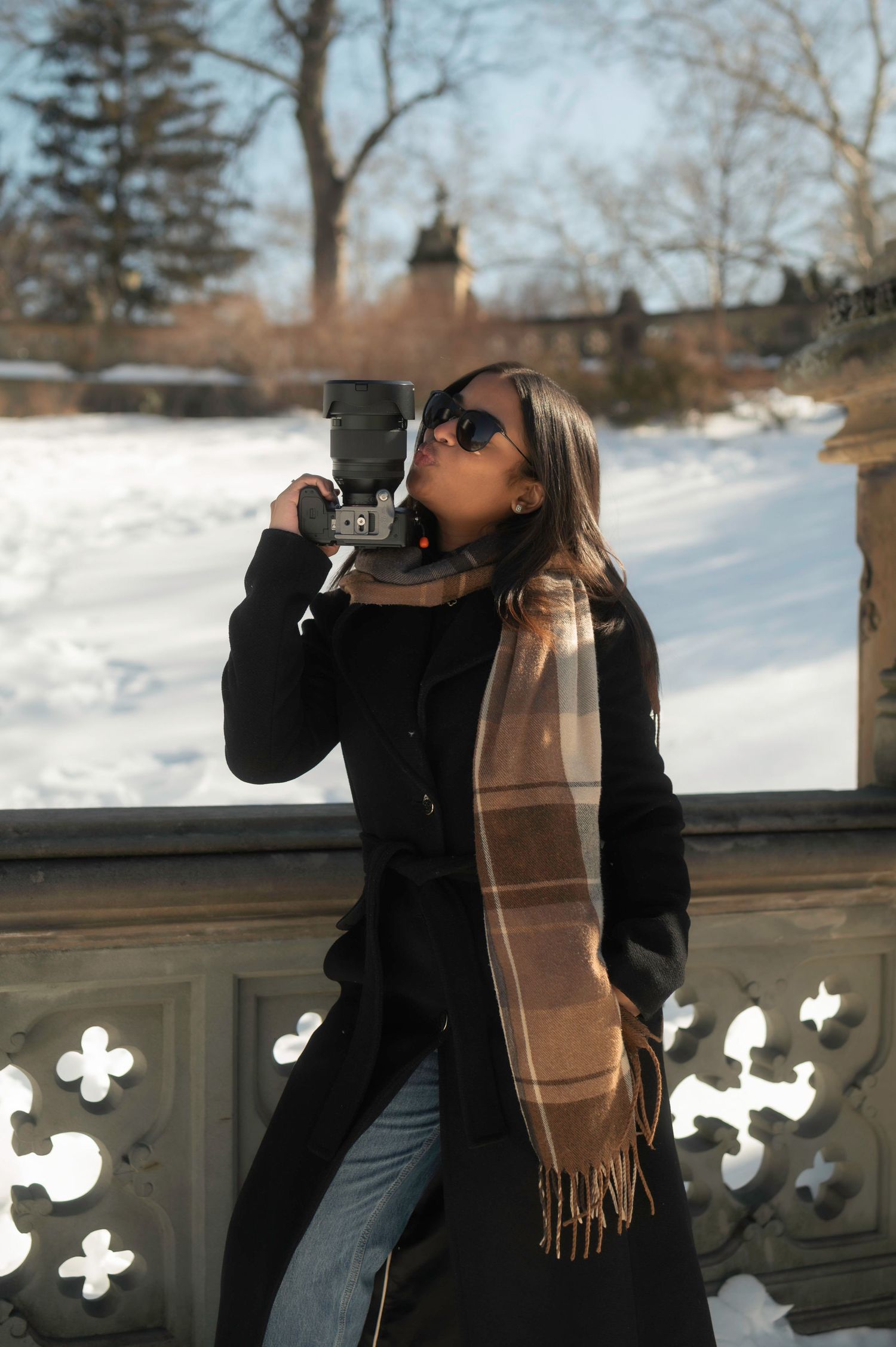 Person in black coat and tan scarf drinks from a bottle while standing in snowy winter landscape.