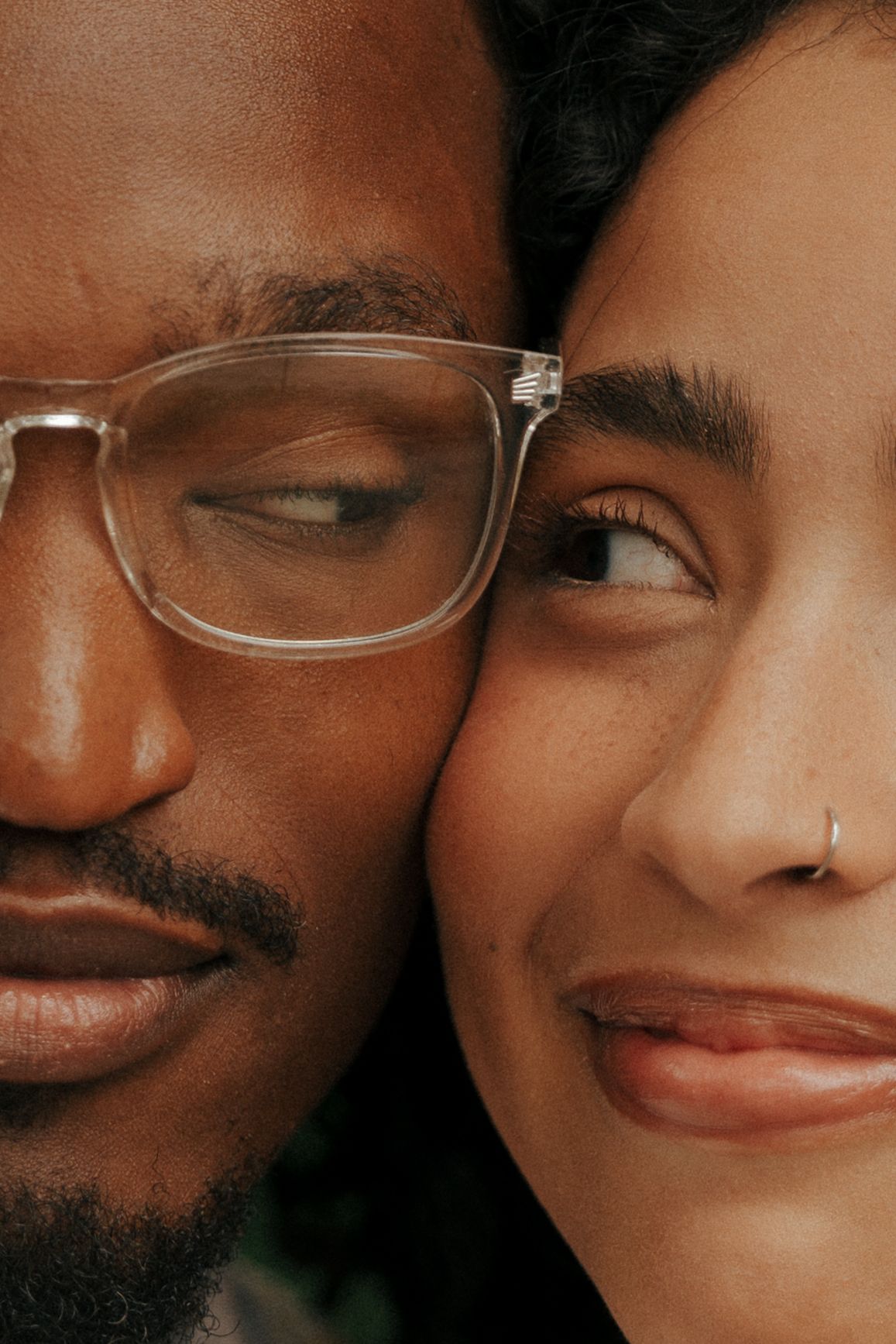 Close-up portrait of a man with clear glasses and a woman with a nose ring, cheeks touching, smiling together.