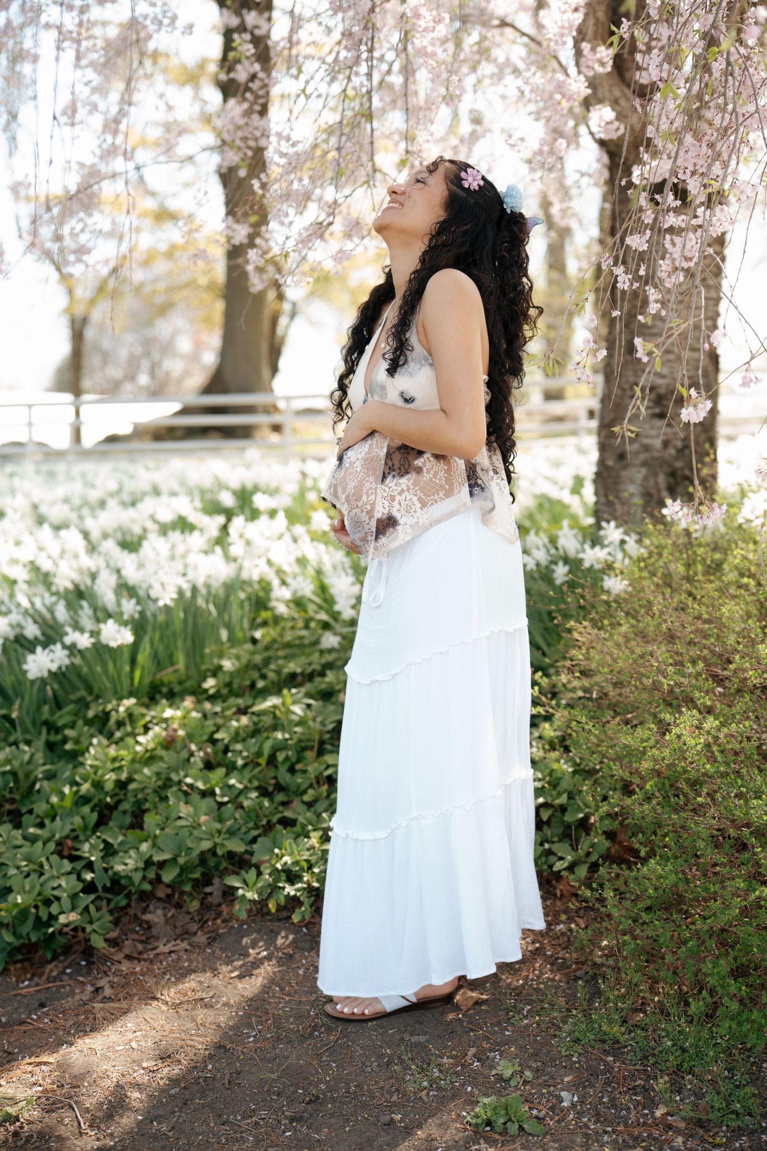A woman in a flowing white dress stands among blooming spring flowers and trees in a dreamy outdoor setting.