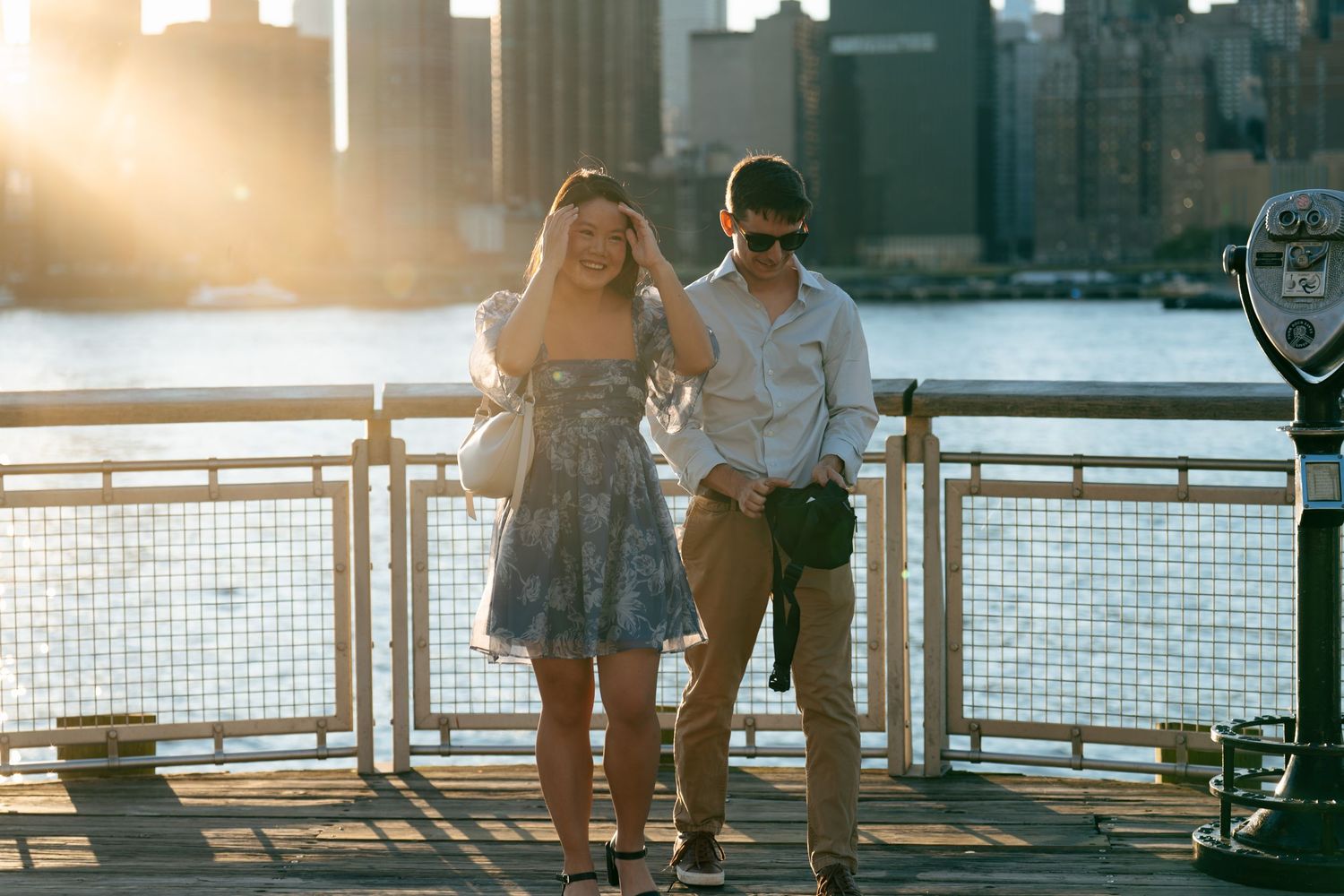 A couple walks along a waterfront railing at sunset with the city skyline in the background.
