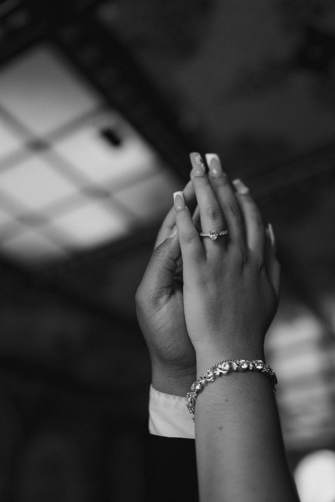 Close-up of clasped hands showing an engagement ring and diamond bracelet in black and white.