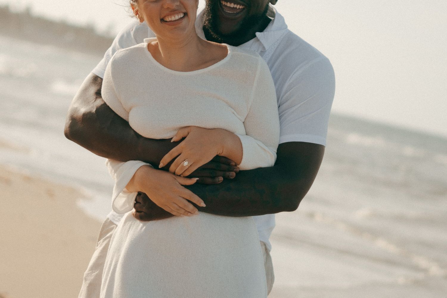Happy interracial couple embracing on a sunny beach, wearing white outfits, sharing a joyful moment together.