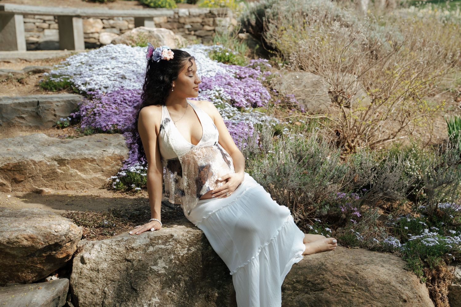 A woman in a white dress rests on stone steps surrounded by purple flowers in a garden setting.