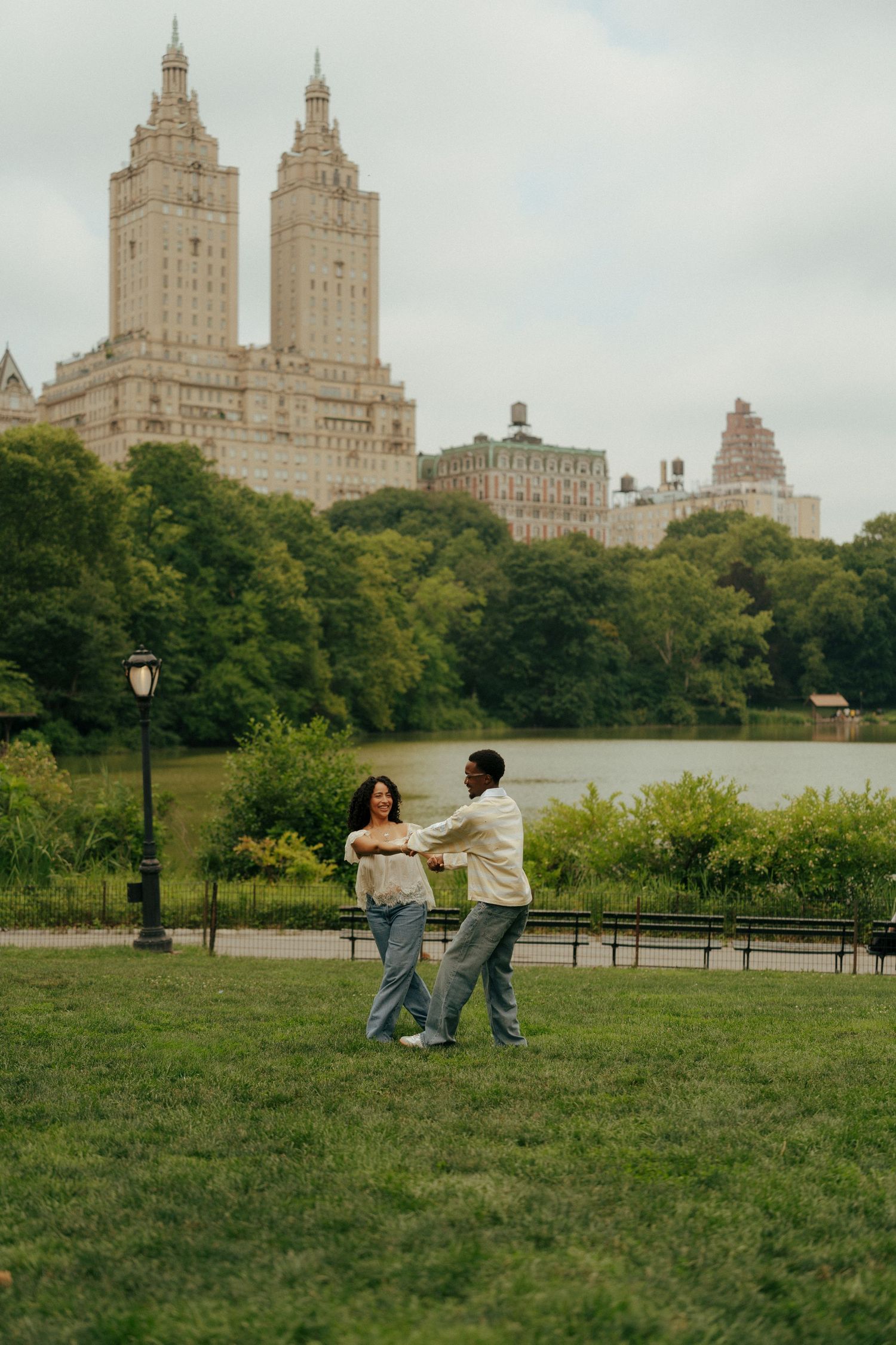 A couple dances joyfully on the grass in Central Park with a lake and city skyline in the background.