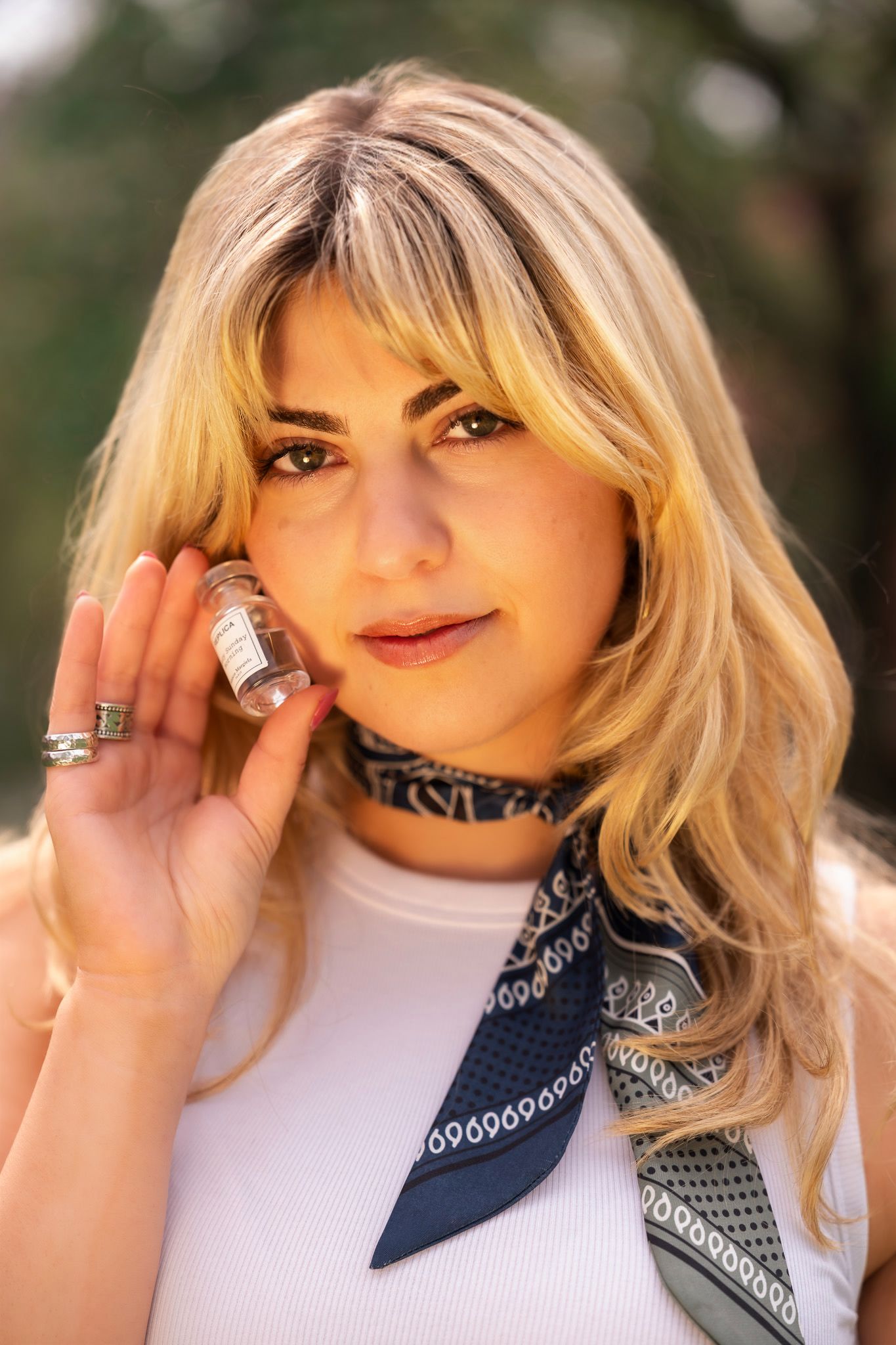 Blonde woman in white top and navy bandana scarf holding a small perfume bottle outdoors.