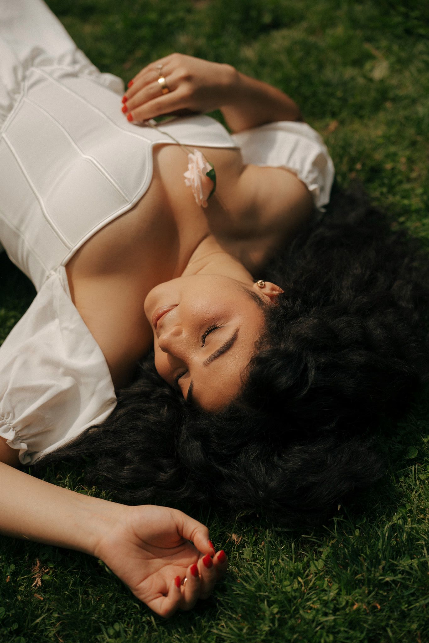 Woman with dark curly hair lying on green grass wearing a white dress with red nail polish.