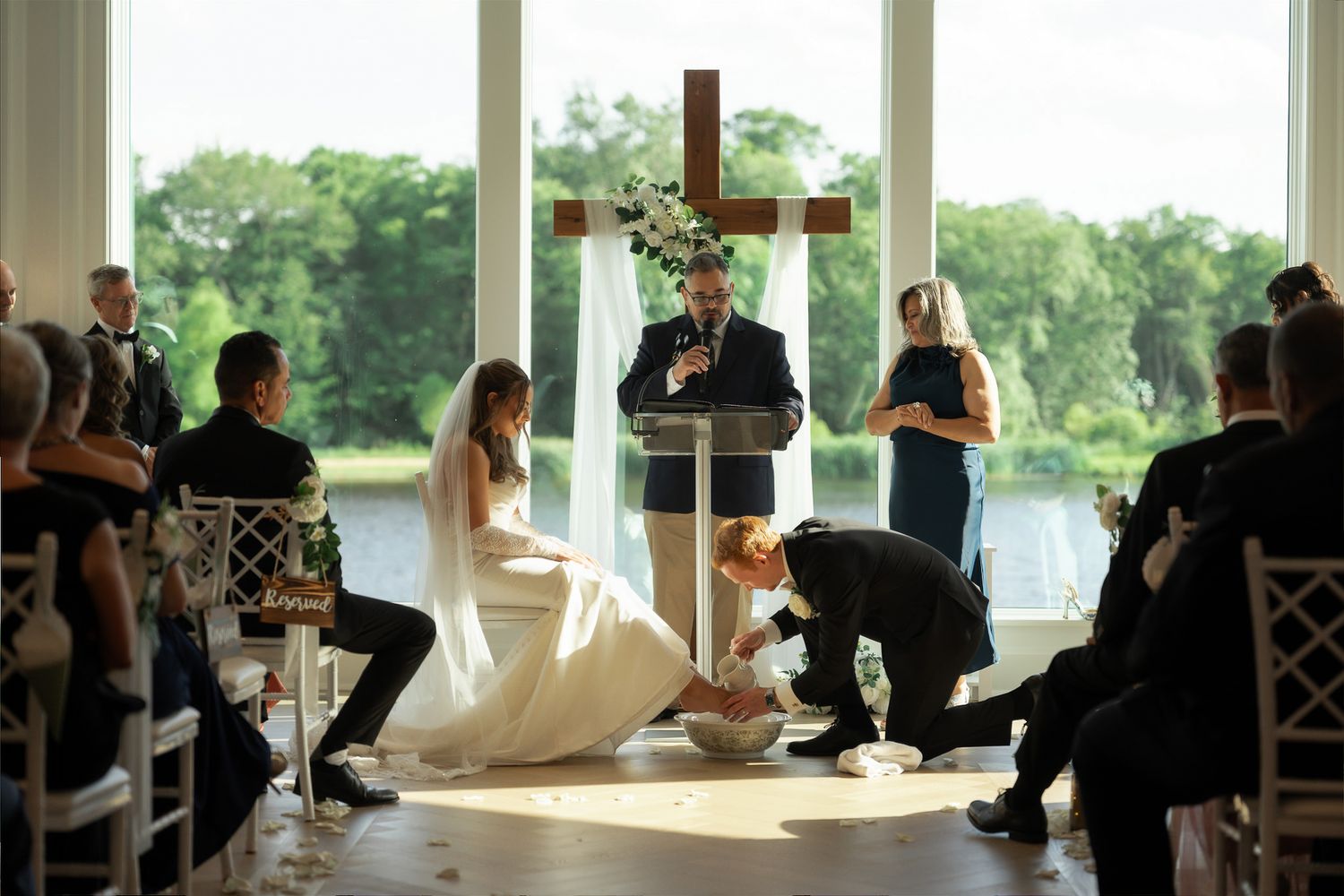 Elegant wedding ceremony in a glass chapel with a cross, officiant at podium, bride in white gown, and groom kneeling.