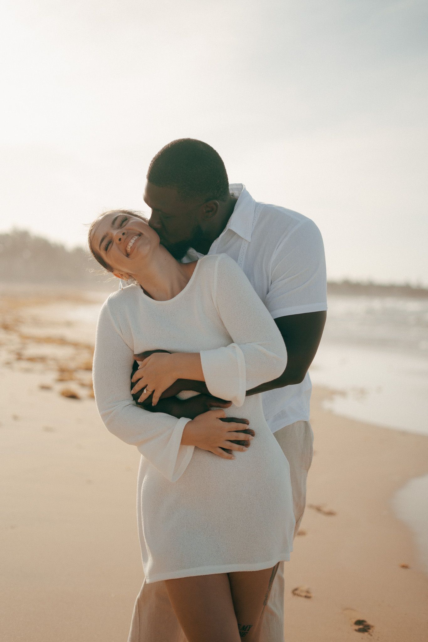 A couple embraces on a sunny beach, the man kissing the smiling woman in a white dress.
