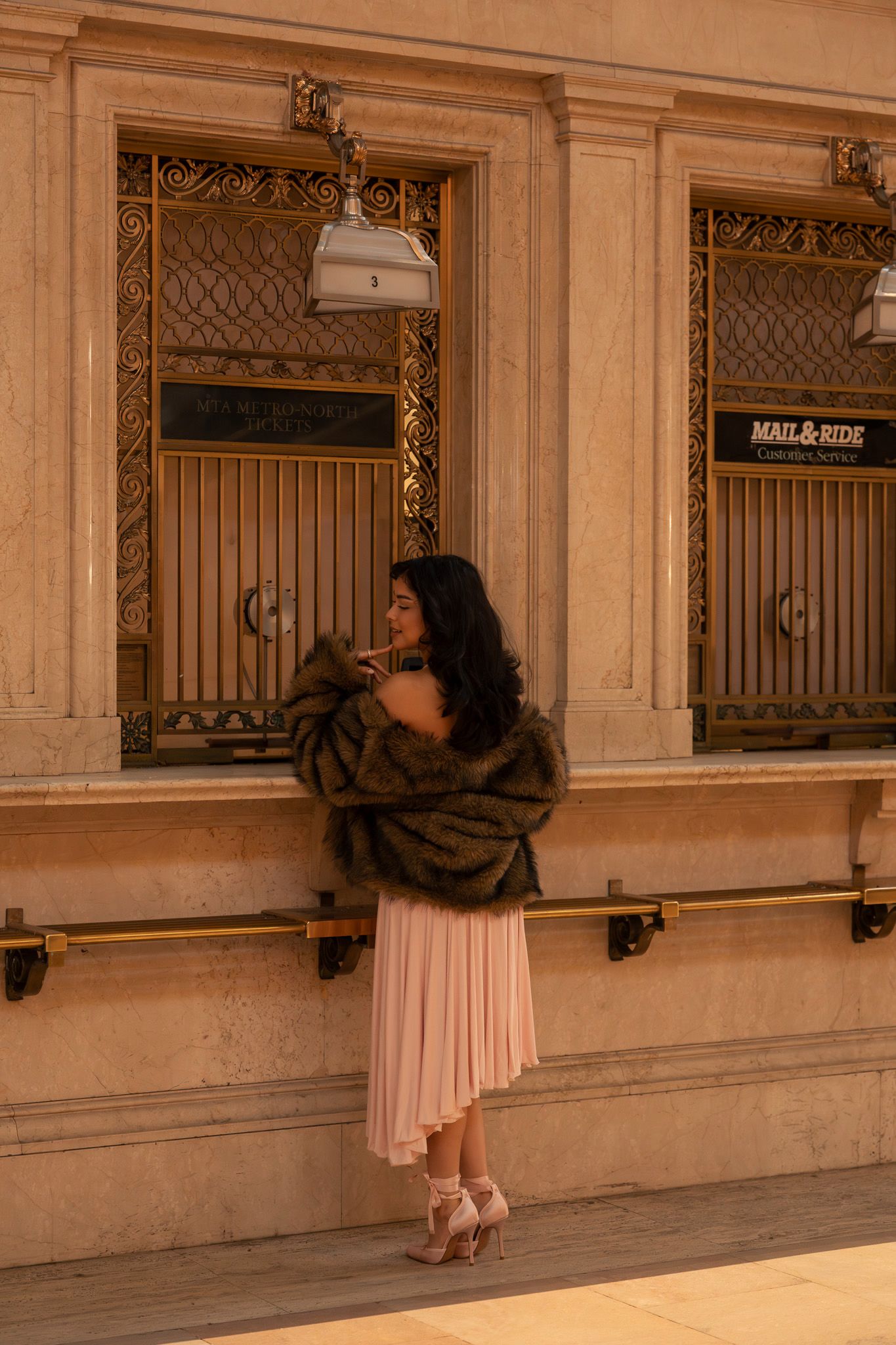 A woman in a pink dress and brown fur coat poses against a beige stone building exterior.