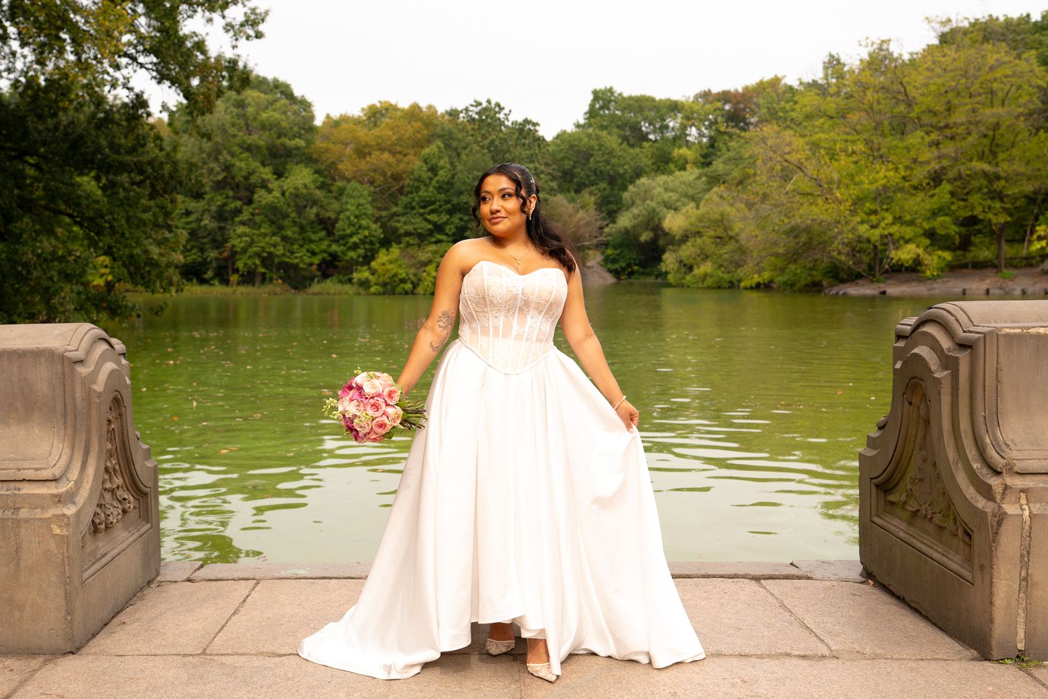 Bride in white wedding gown standing near a lake with fall foliage in background.