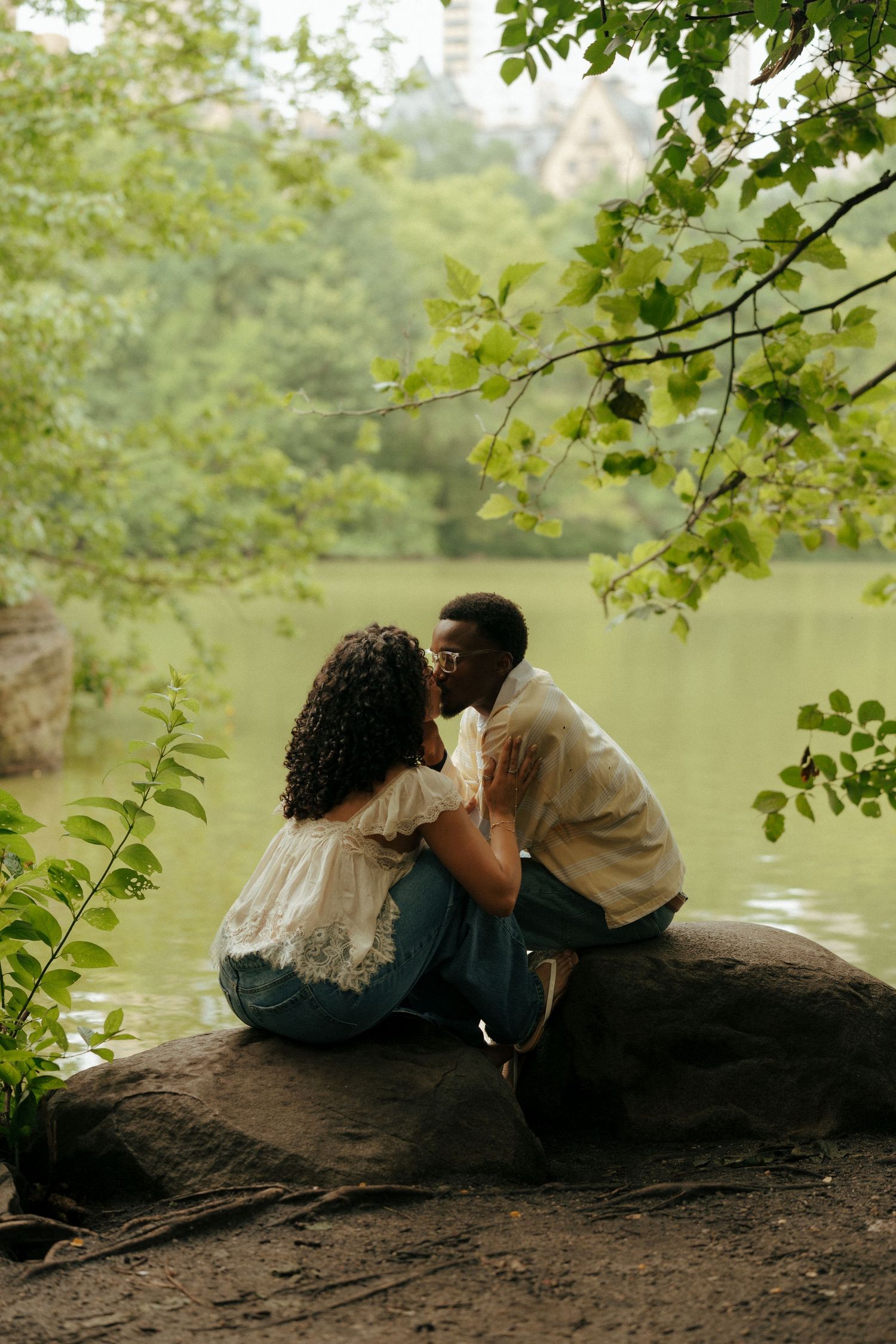 A couple shares a tender kiss while sitting on a rock by a serene green lake in a park.