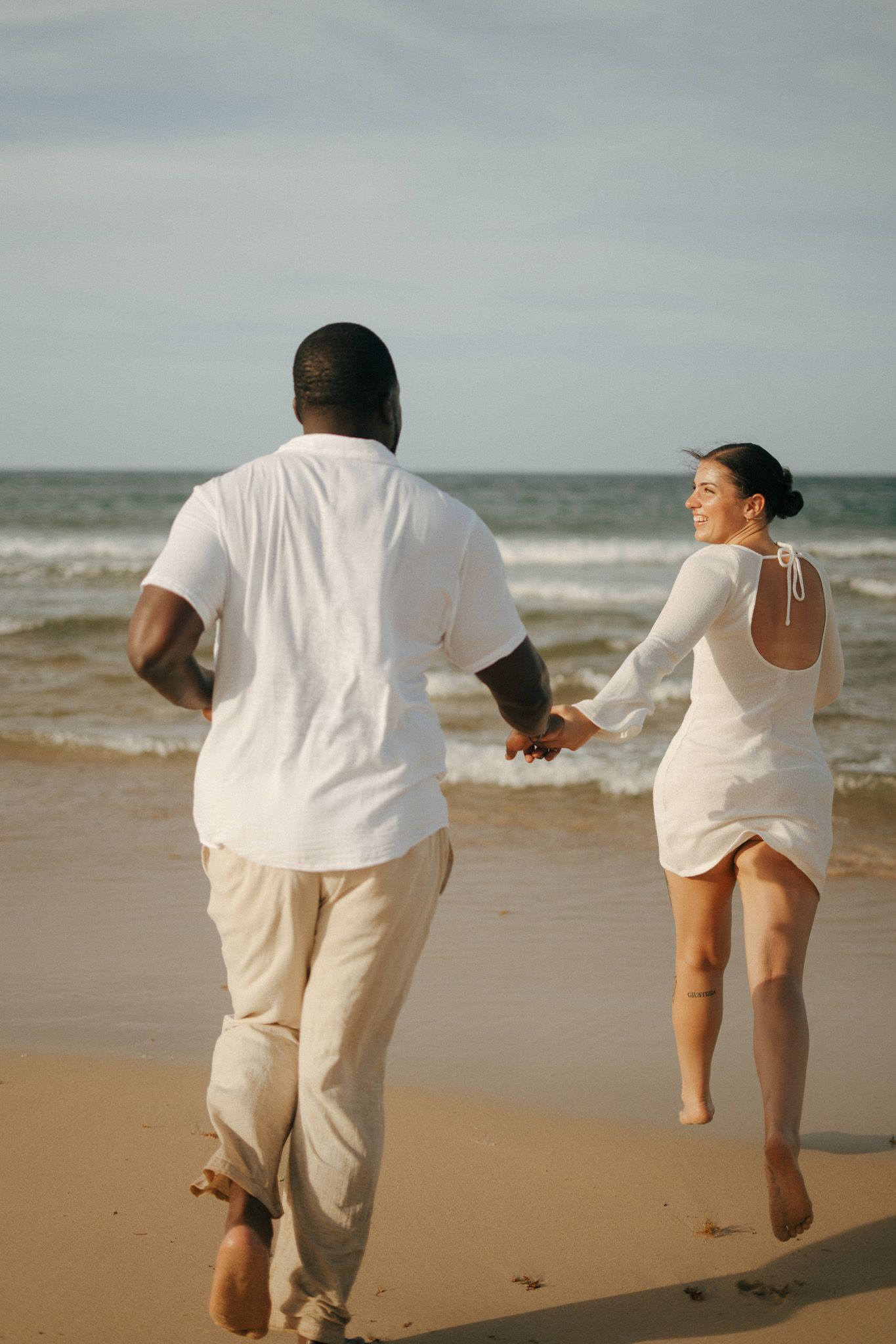 Couple in white outfits holding hands running on a sandy beach toward ocean waves on a sunny day.