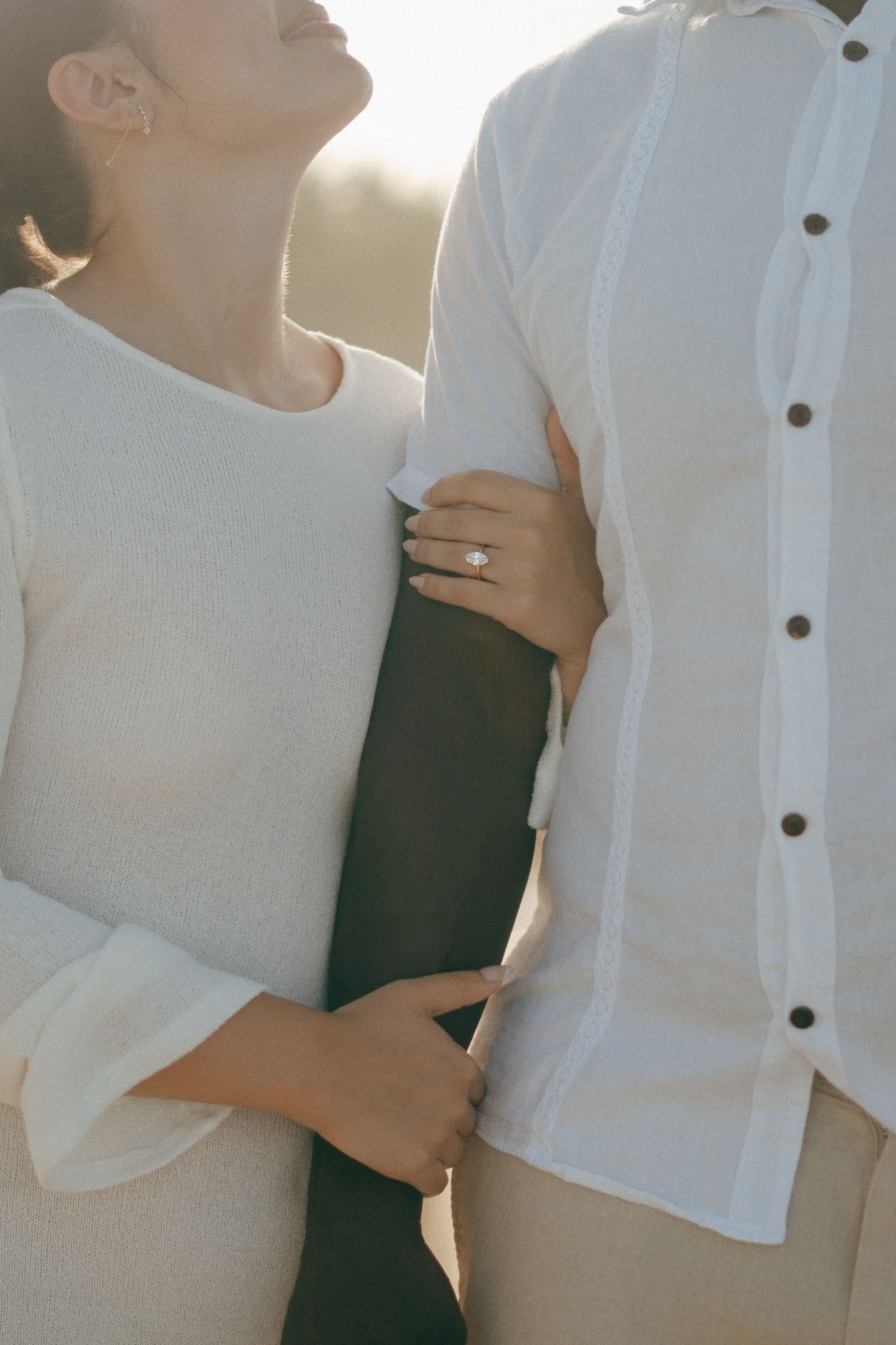 Couple in white outfits embracing outdoors, woman wearing diamond engagement ring, soft backlit natural light.