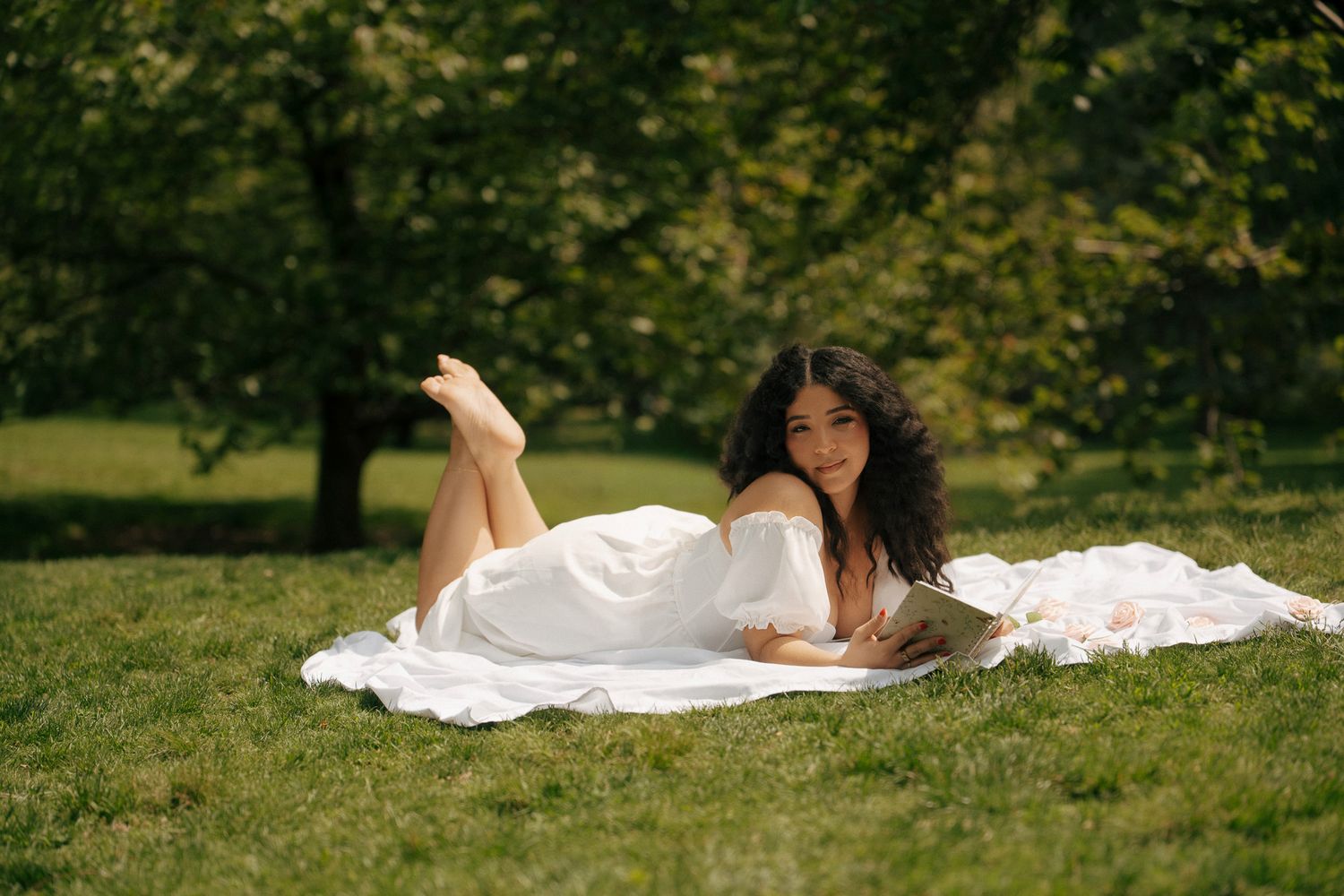 Young woman in white dress lying on blanket in sunny green park, smiling at camera with legs raised.