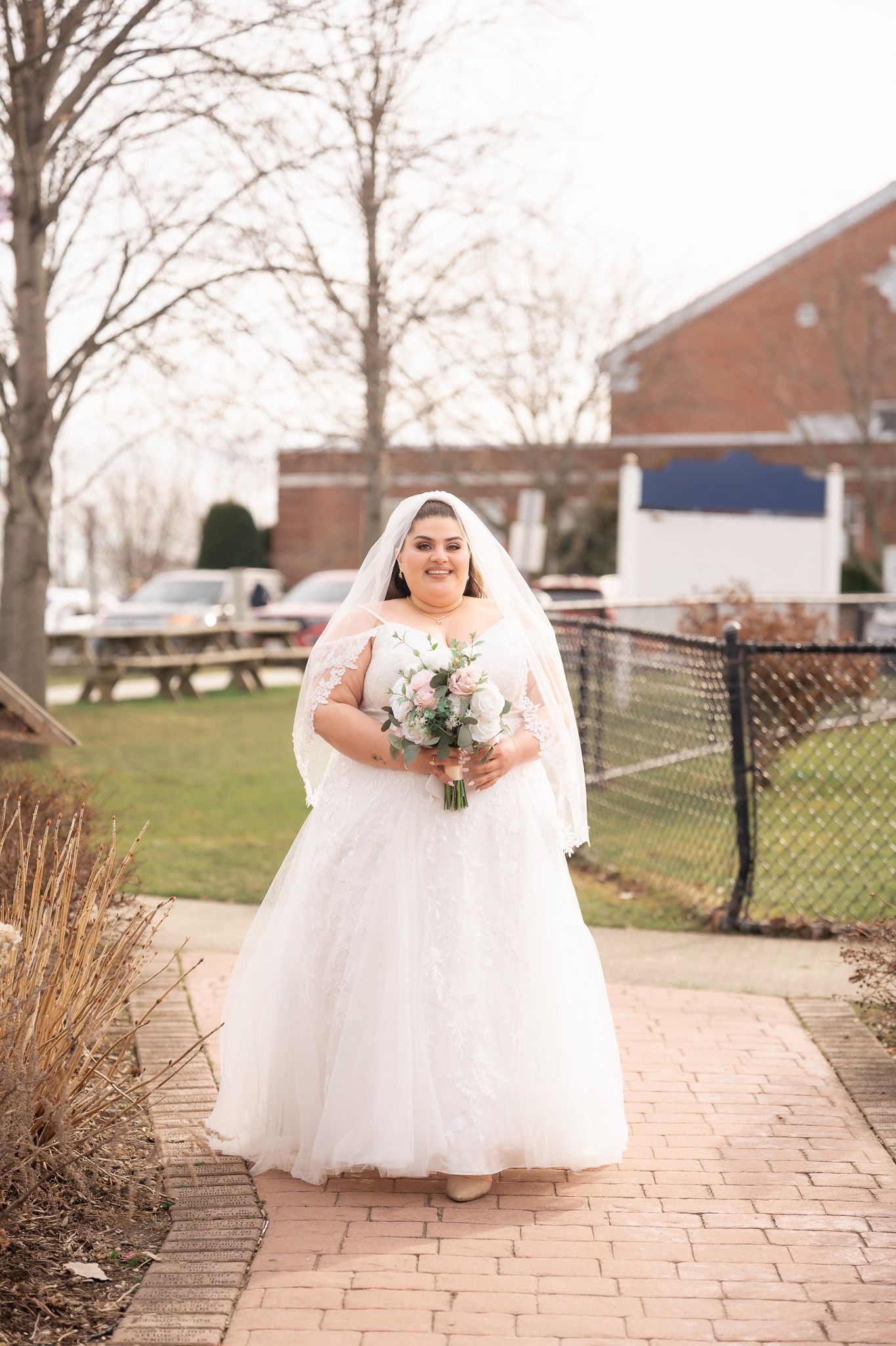 Bride in white ball gown and veil holding bouquet walks on brick path outdoors on a cloudy winter wedding day.