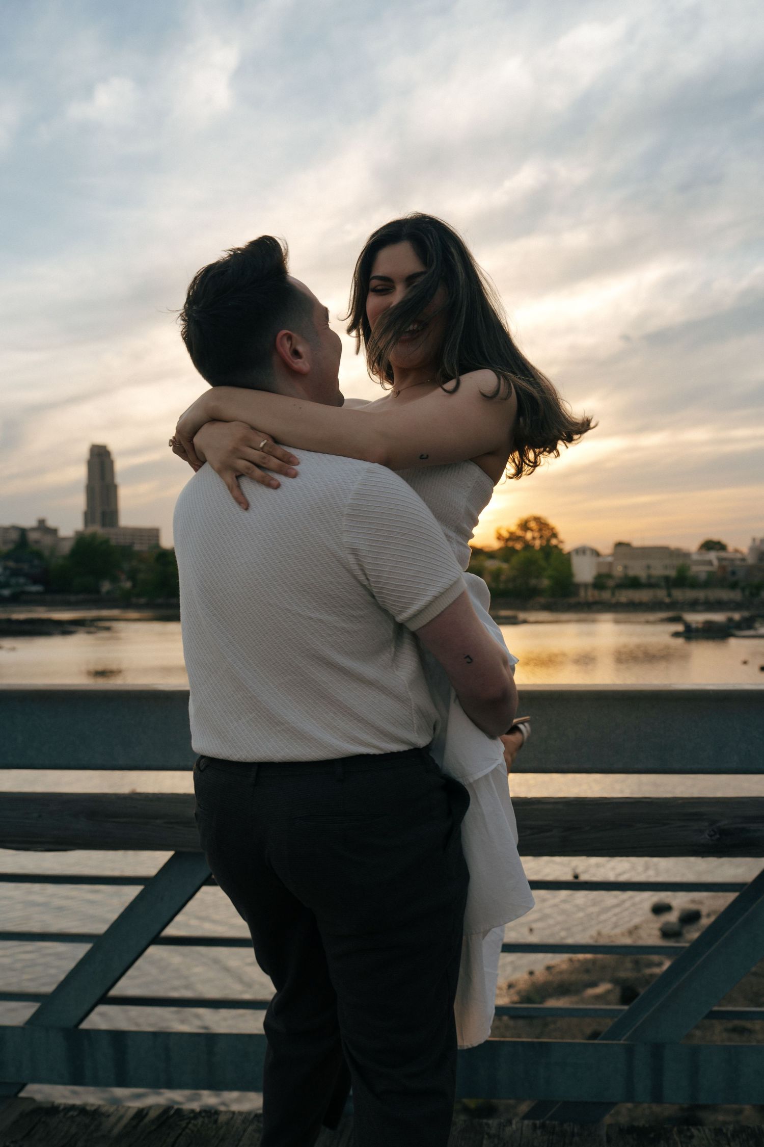 Romantic waterfront photoshoot with urban skyline silhouetted against cloudy sunset.