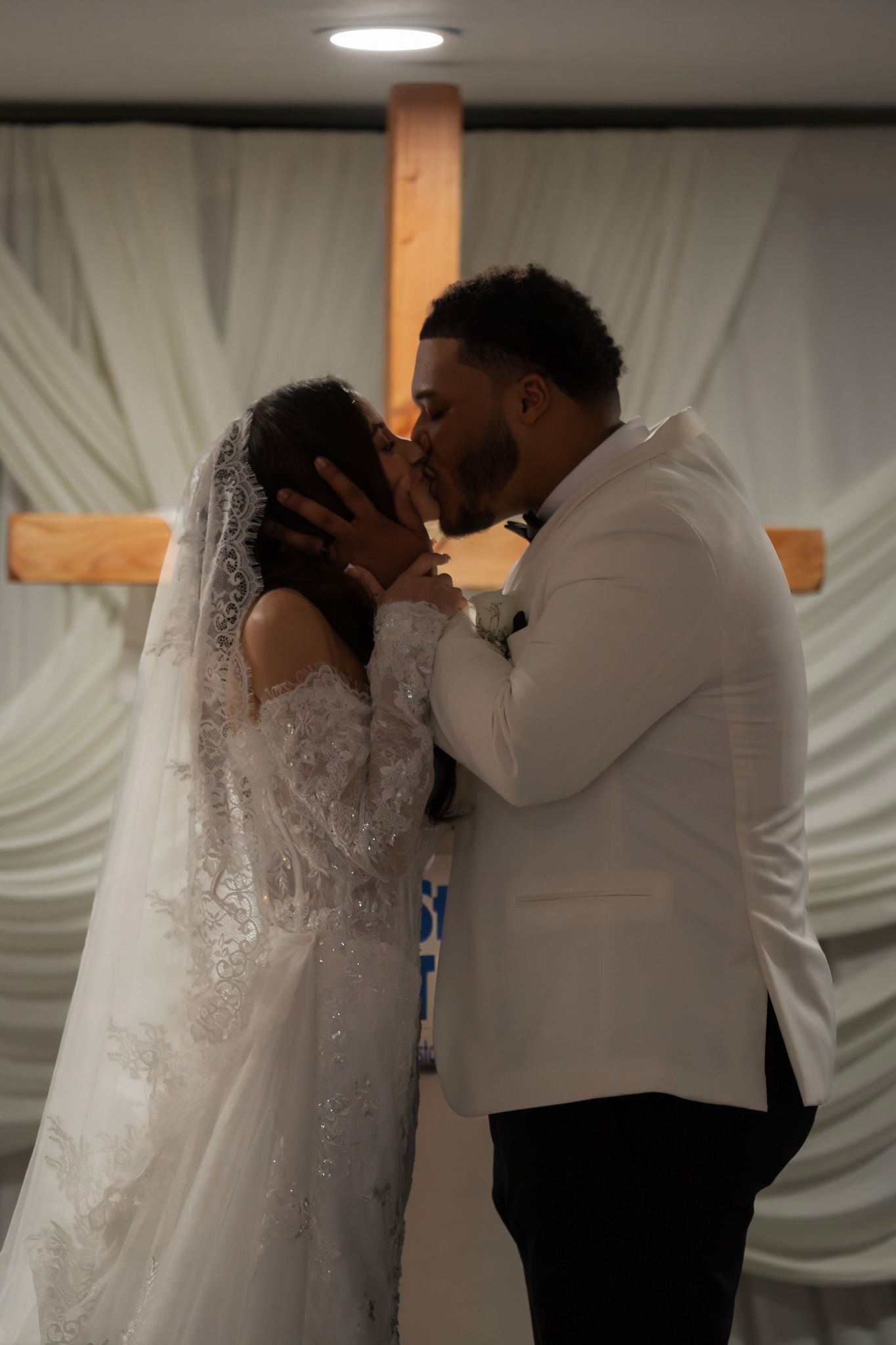 Newlyweds share their first kiss during an intimate wedding ceremony in front of a white draped backdrop.