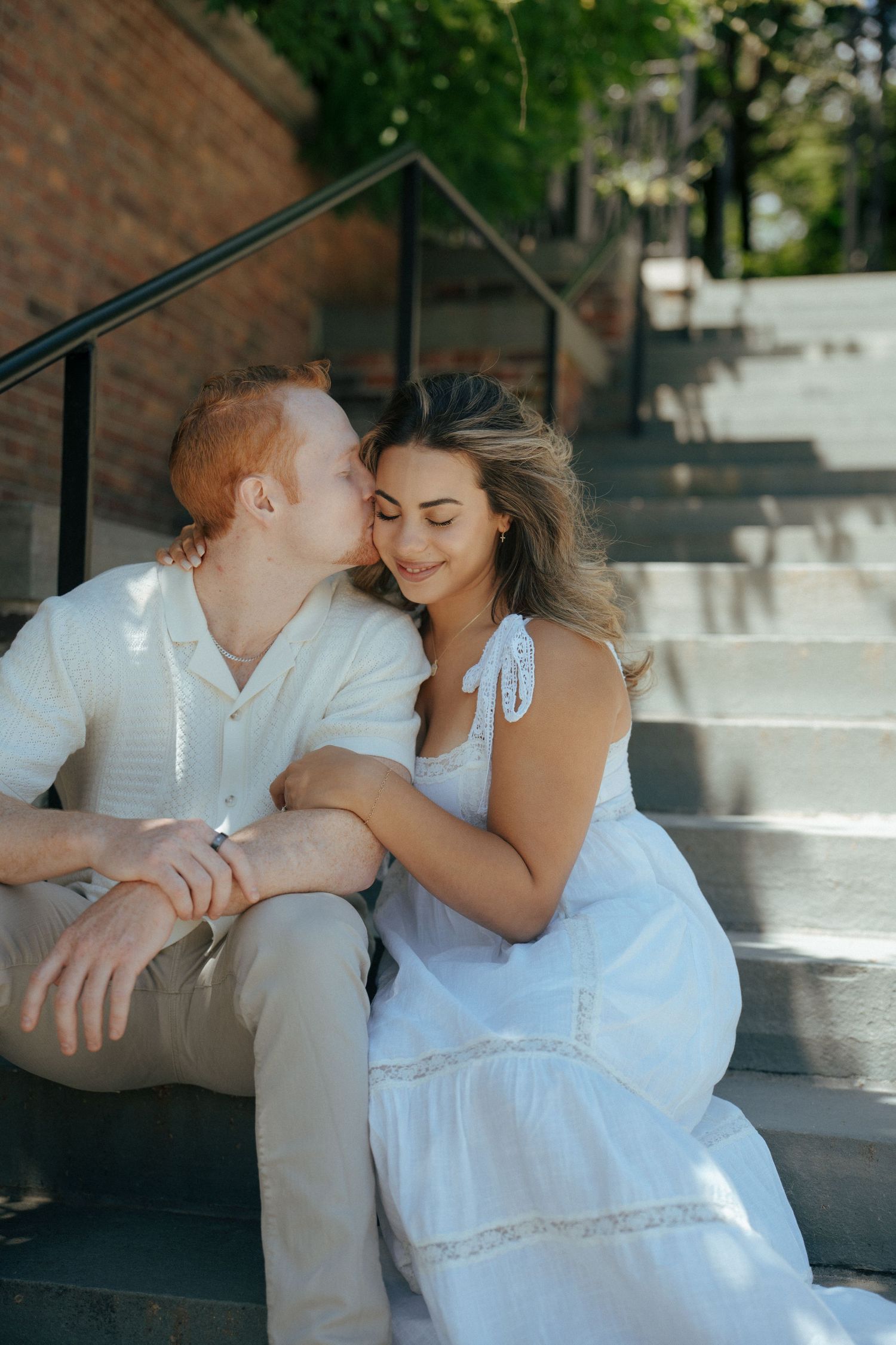 A couple shares an intimate moment while sitting together on concrete steps on a sunny afternoon.