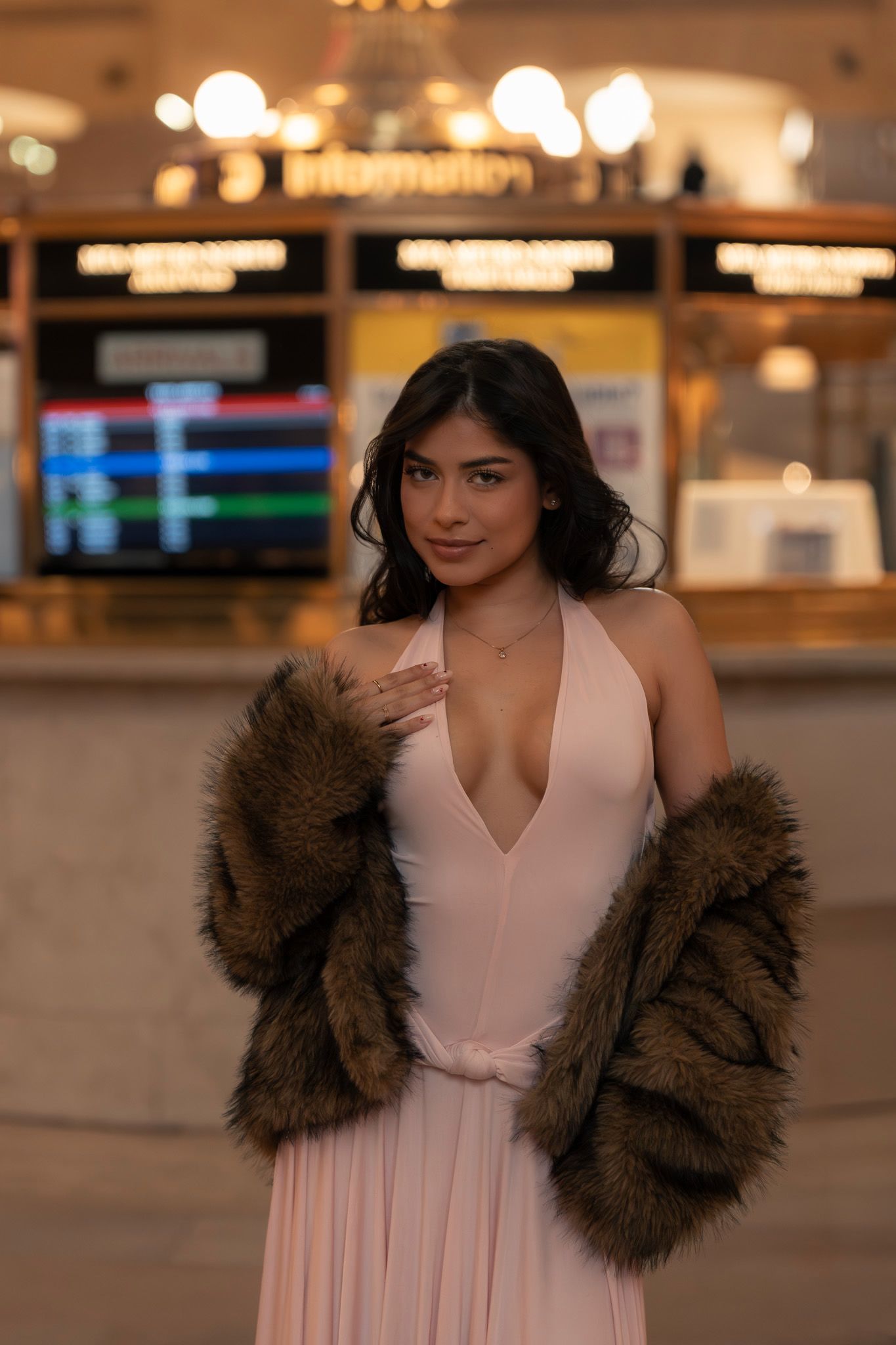 A person in a pink evening gown and brown faux fur coat stands in front of an illuminated storefront display.