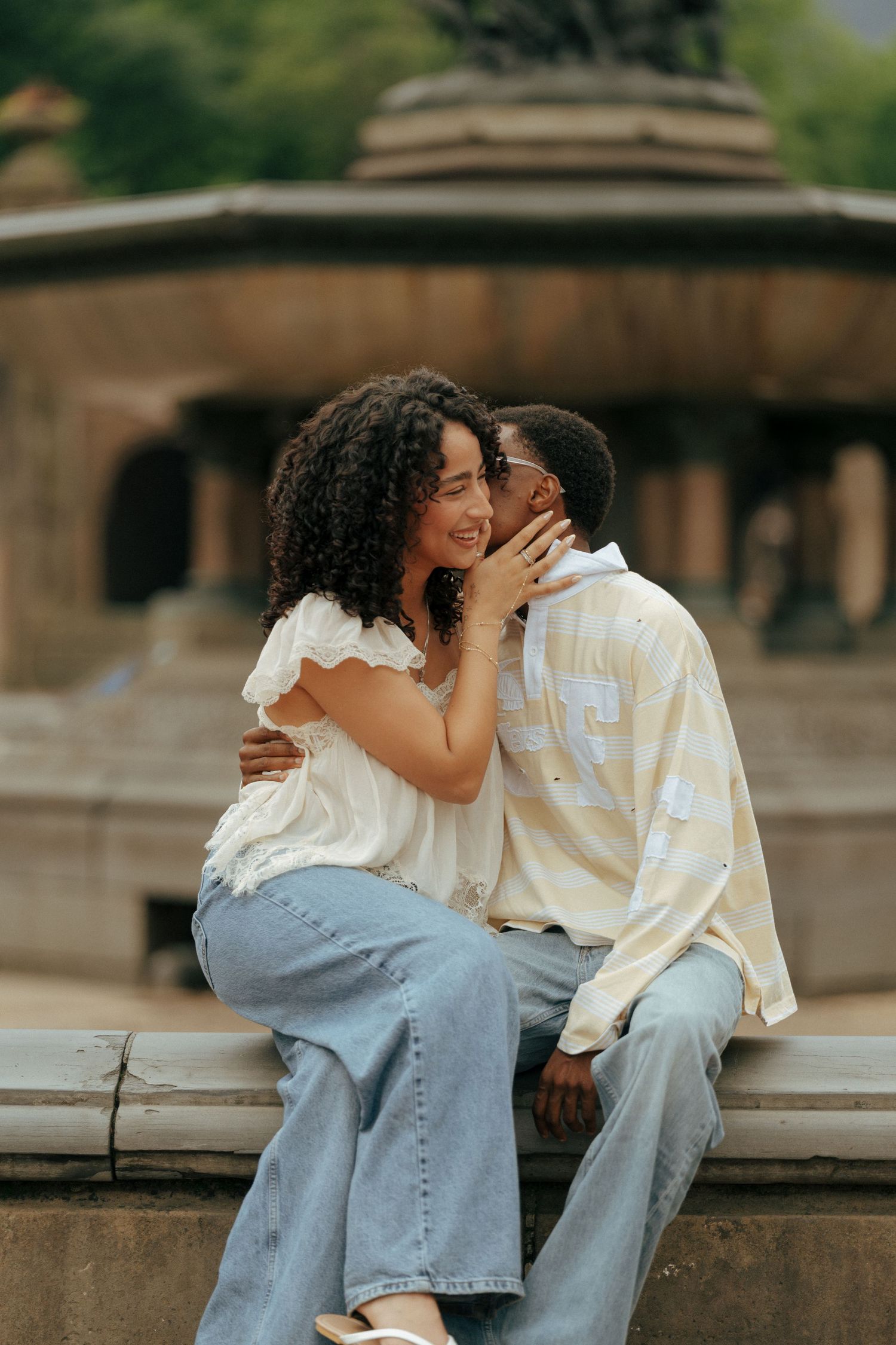 A couple shares a romantic moment sitting on a fountain ledge, with the man kissing the woman's cheek outdoors.