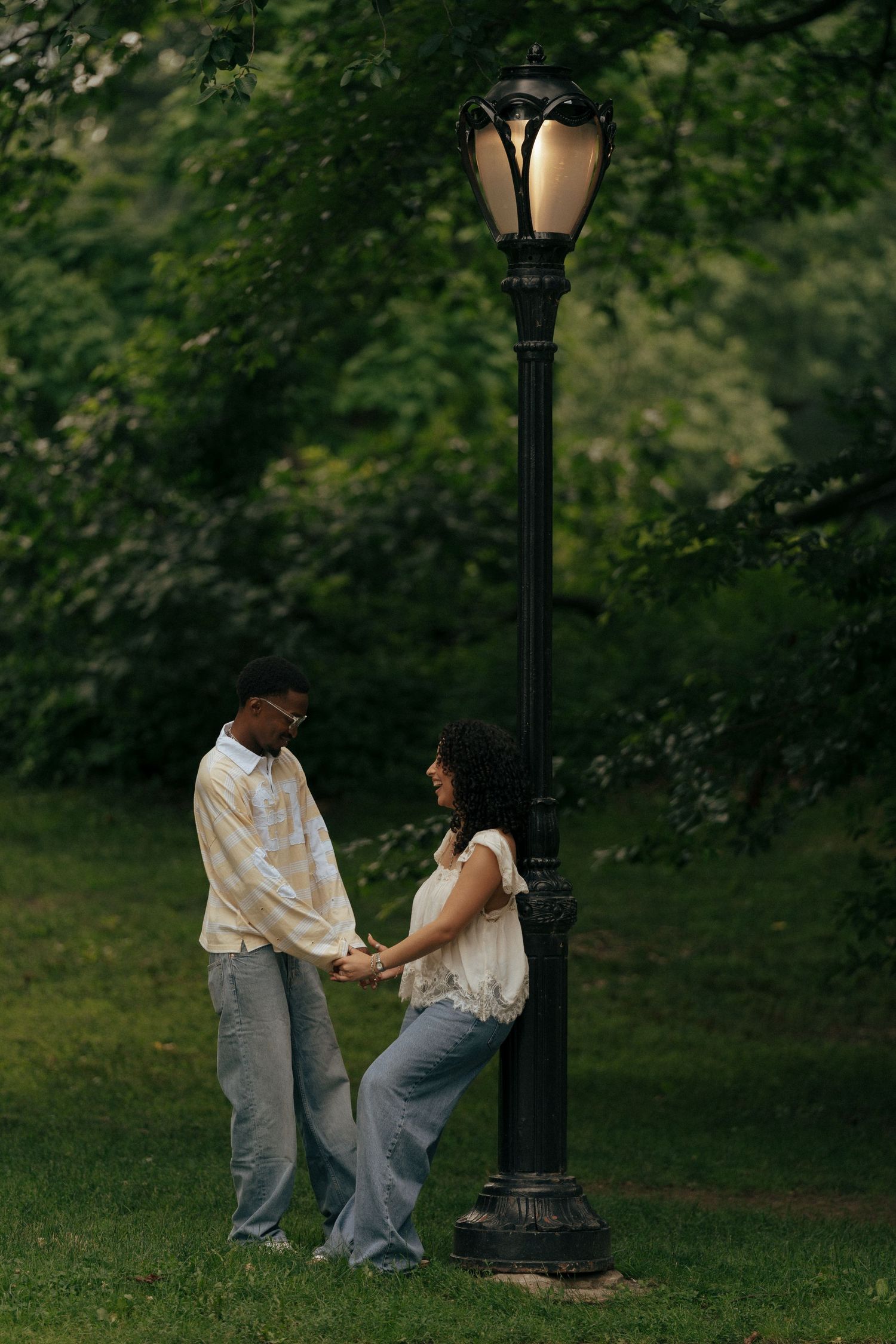 A couple holds hands romantically under a glowing park lamp surrounded by lush green trees.