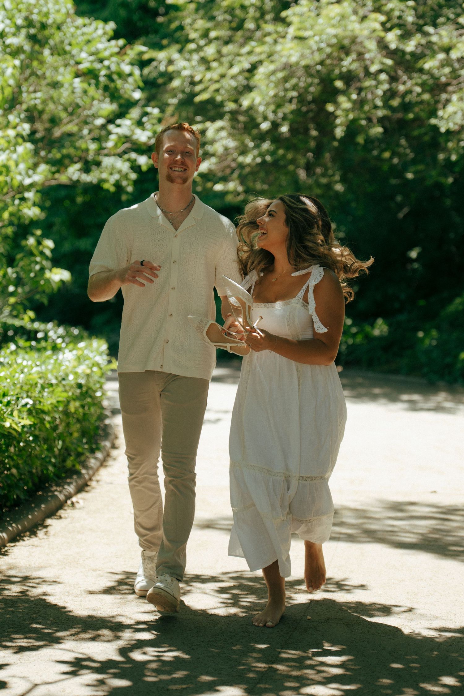 A couple in white outfits walking and laughing together on a sunny garden path.