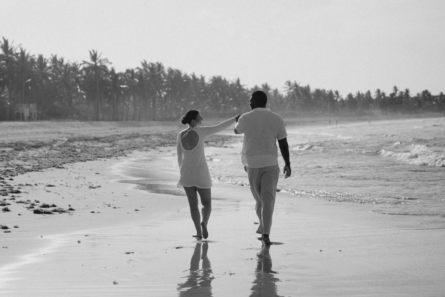A couple walks barefoot along a tropical beach shoreline in black and white photography.