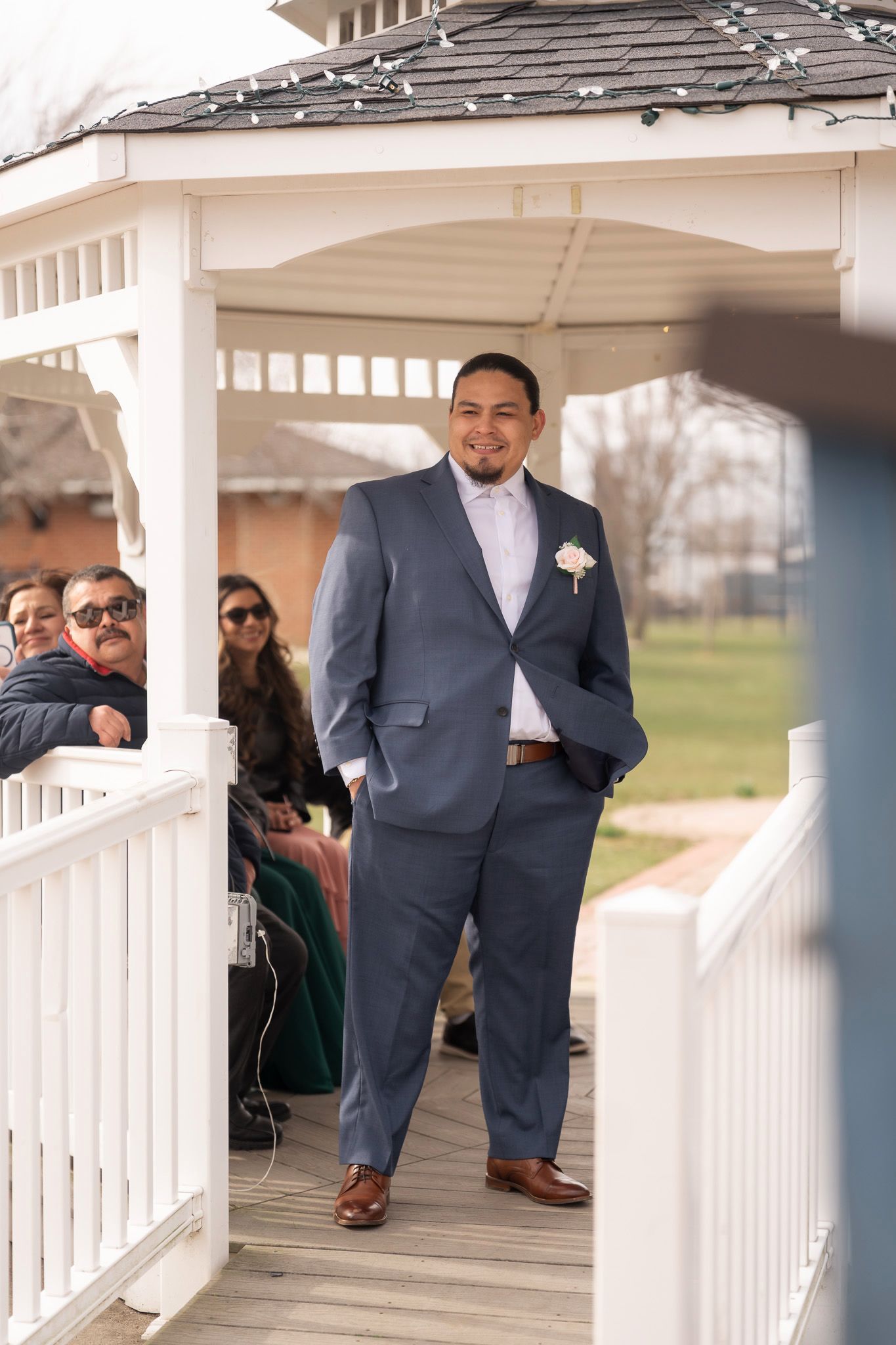 Groom in blue suit with boutonniere stands in white gazebo at outdoor wedding ceremony.