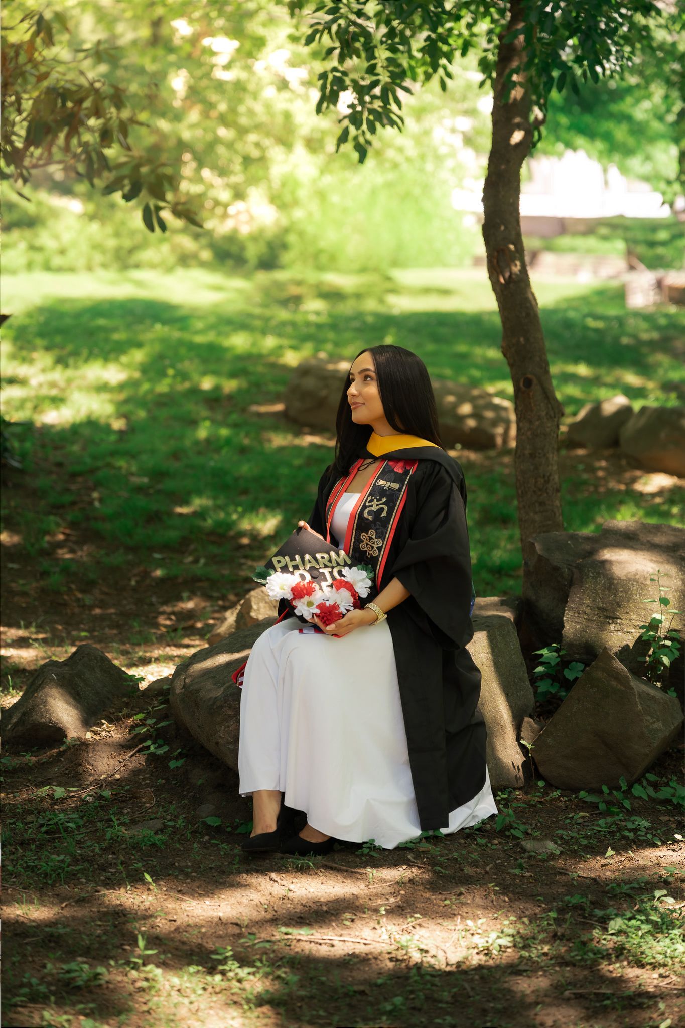 Graduate in black cap and gown holding flowers, seated on rocks in a lush green park setting.