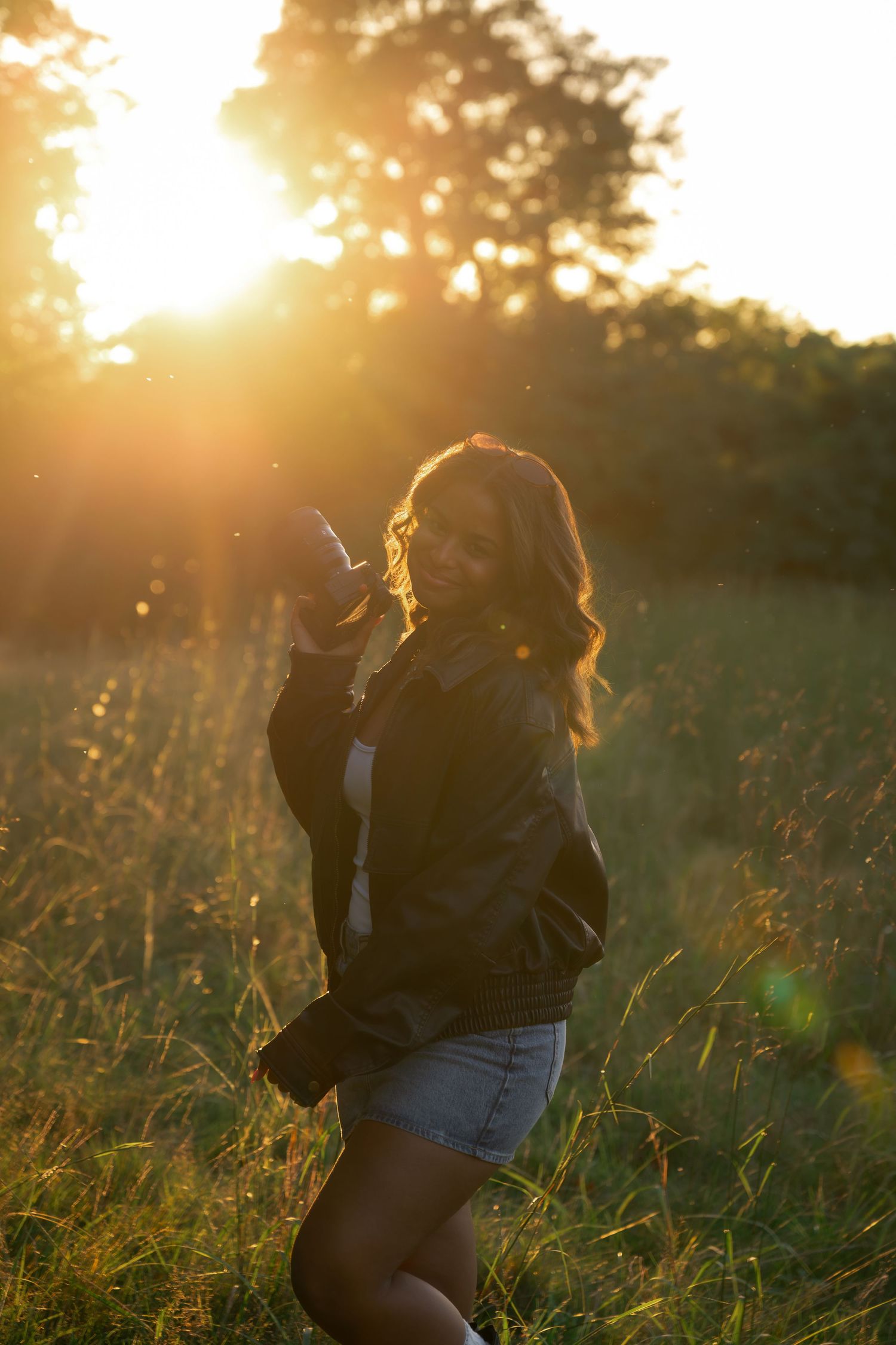 Young woman in black jacket and denim shorts standing in golden sunset field with warm glowing backlight.
