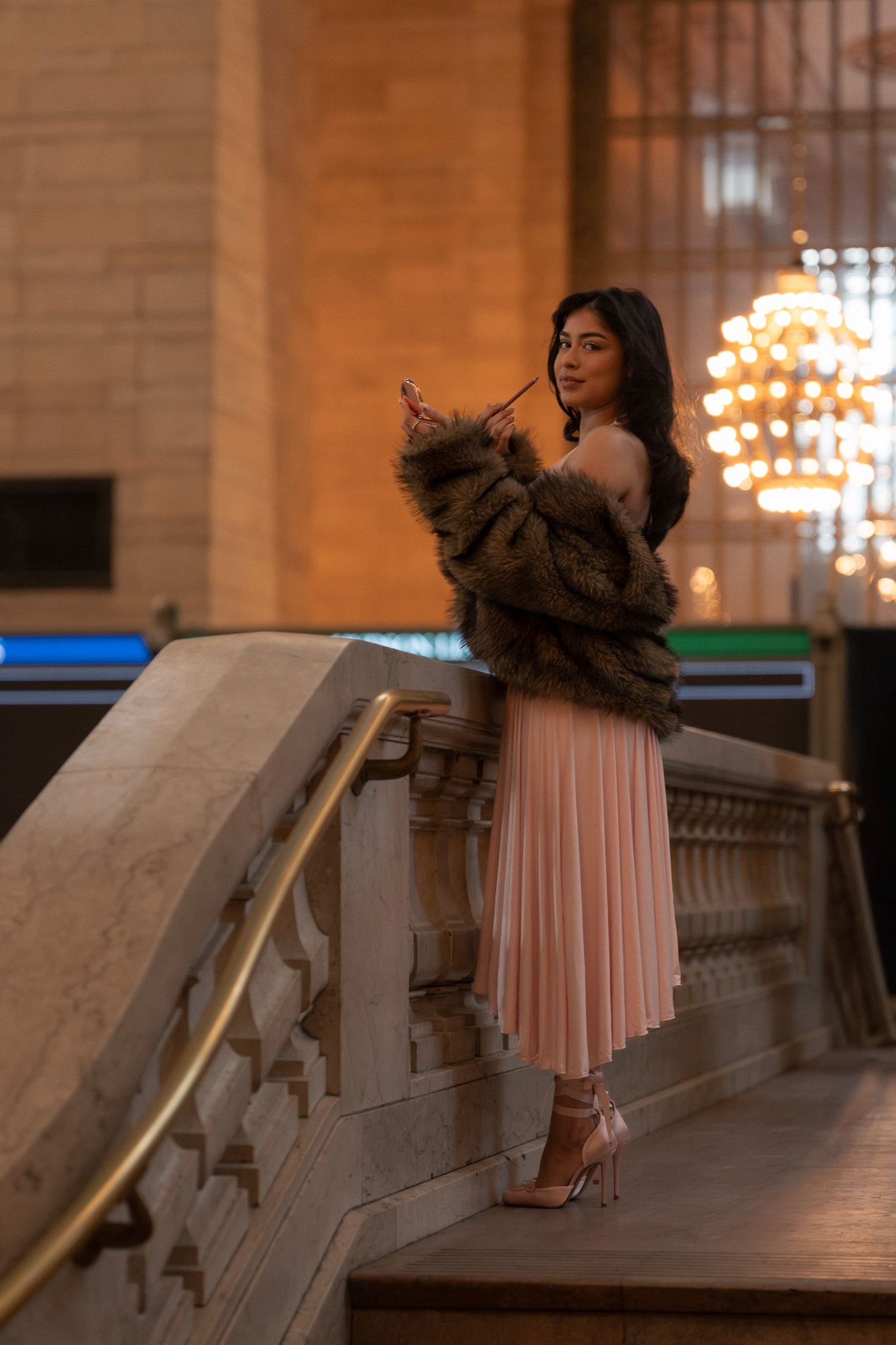 Elegant woman in pink pleated skirt and fur coat poses on marble staircase with chandelier in background.