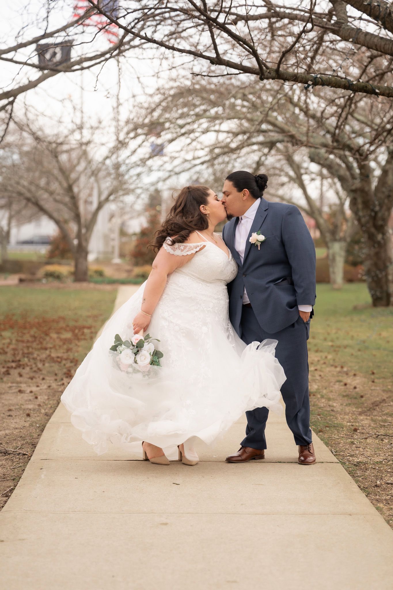 Two brides share a romantic dip kiss on a garden path, one in a white gown holding roses, the other in a blue suit.