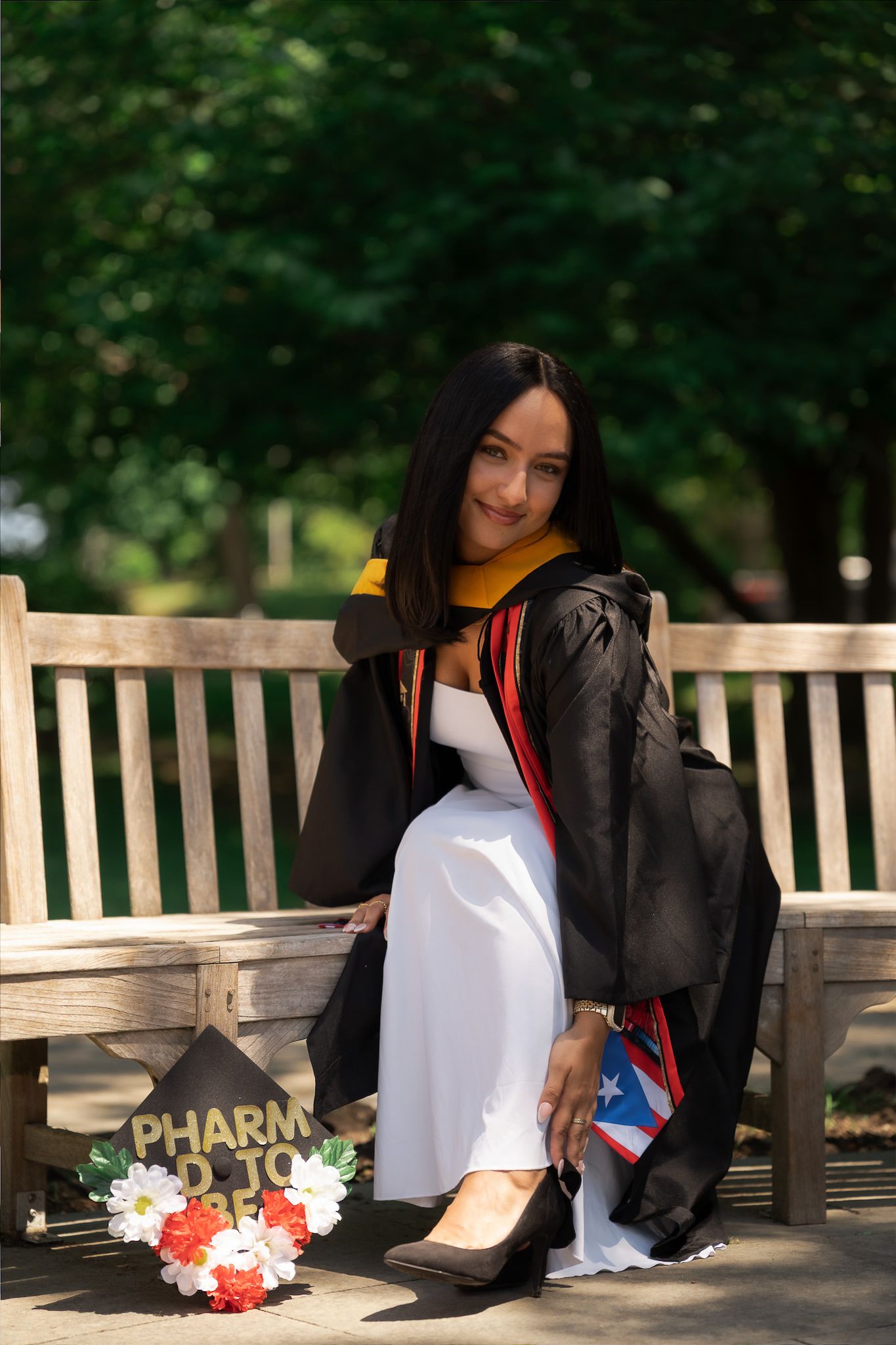 Young woman in black graduation gown sits on bench with PharmD cap and Puerto Rican flag, celebrating pharmacy school graduation.