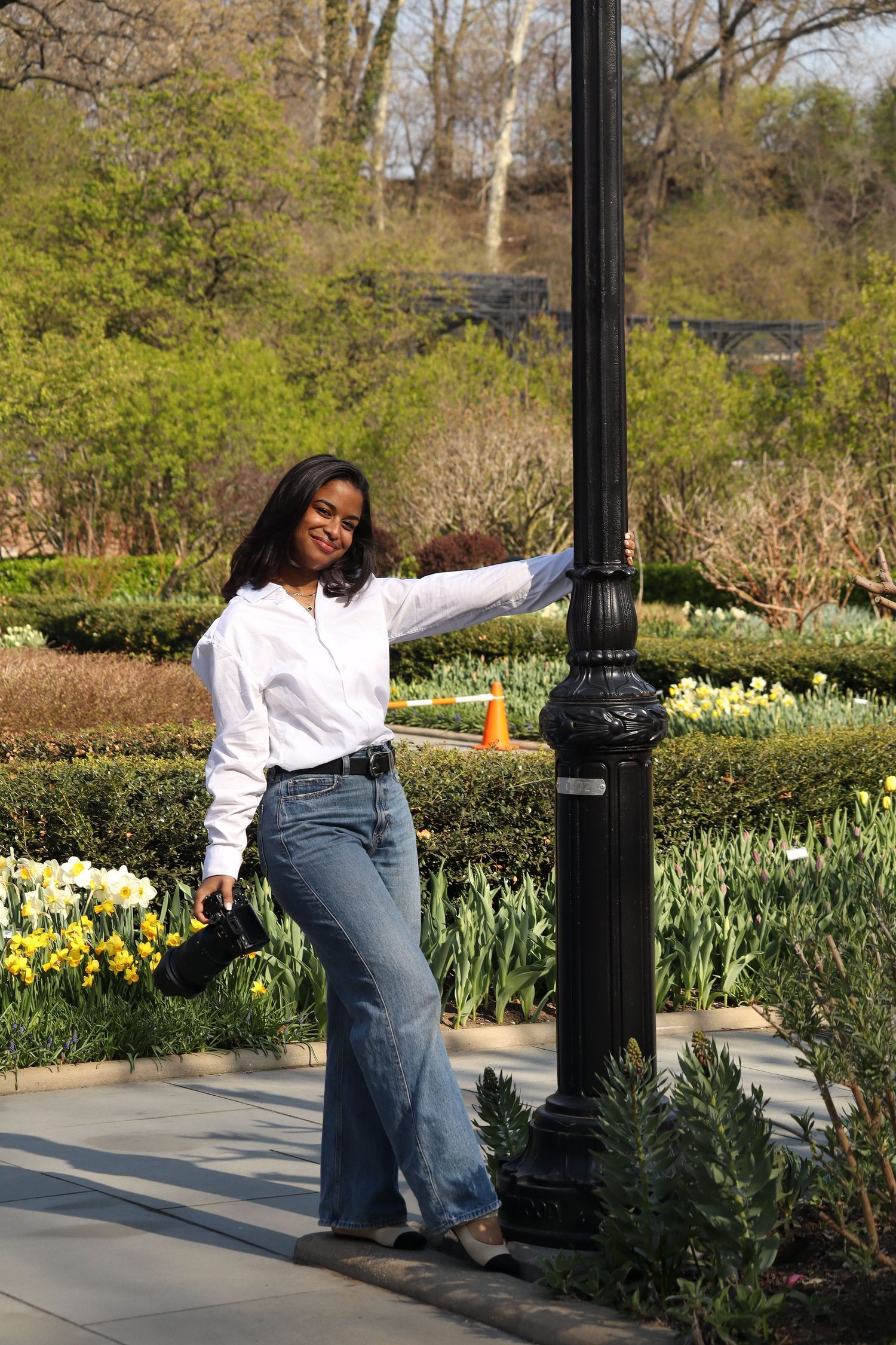 A smiling woman in white shirt and flared jeans poses by a black lamp post in a blooming garden park.