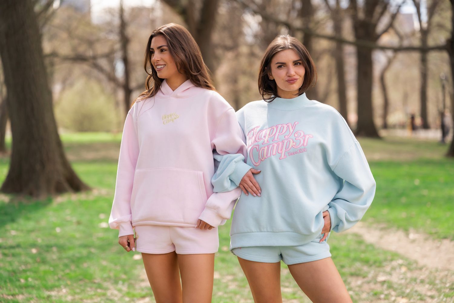 Two women wearing pastel pink and blue matching sweatshirt sets posing outdoors in a park setting.