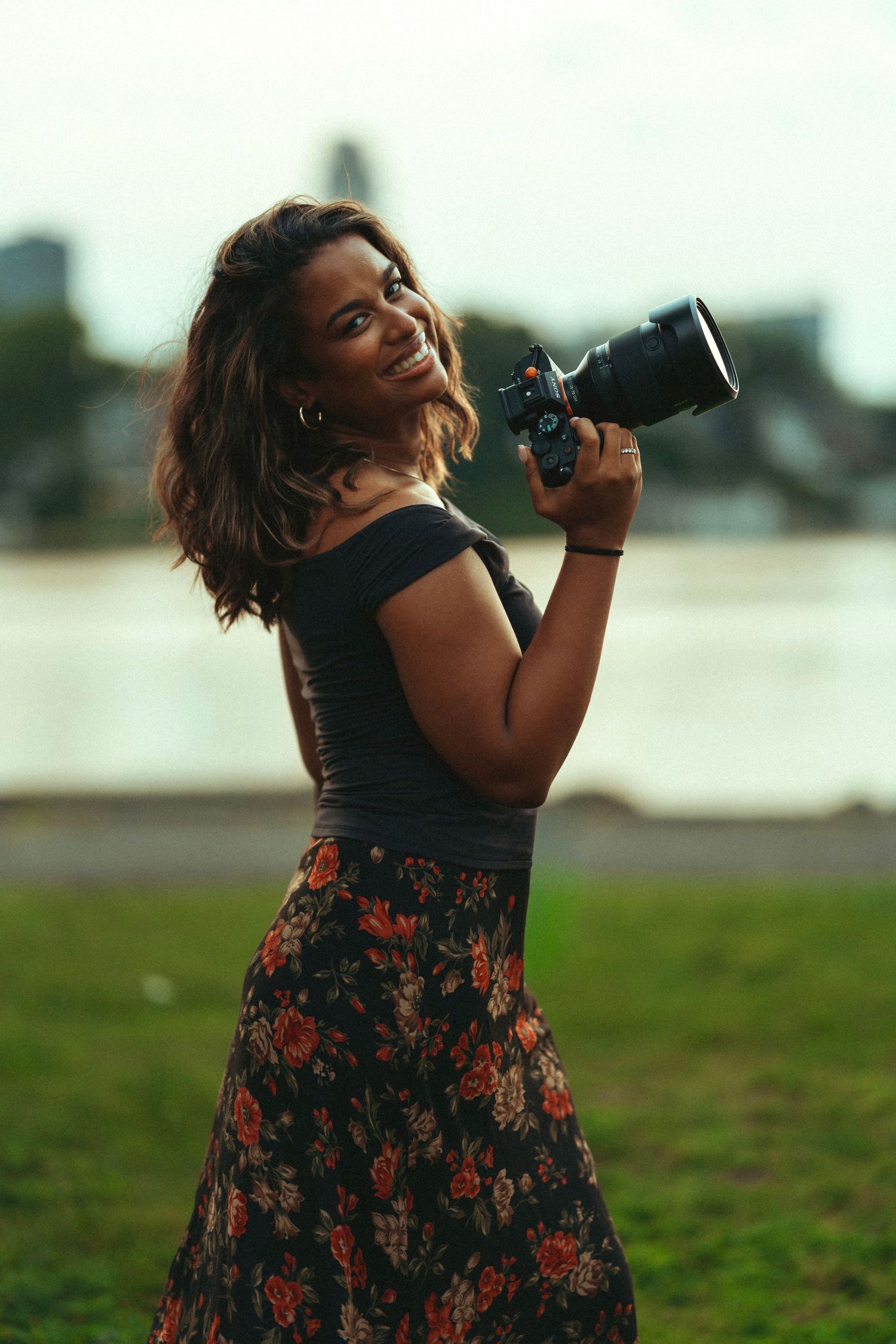 Smiling woman holding a DSLR camera with telephoto lens, wearing floral skirt outdoors near water.