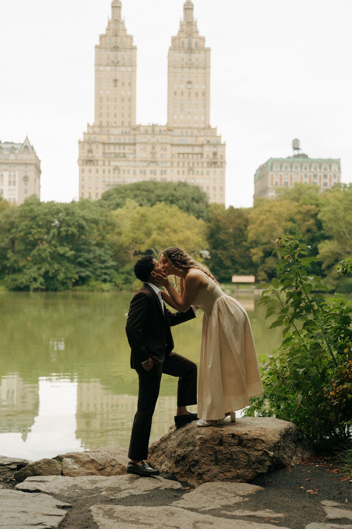 Couple sharing a romantic kiss on rocks by a lake in Central Park, NYC, with the San Remo building in the background.