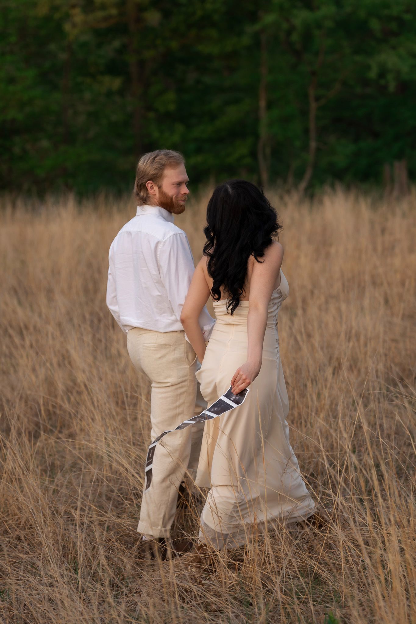 Couple walking hand-in-hand through dry grass field, woman in flowing beige dress holding ultrasound images.